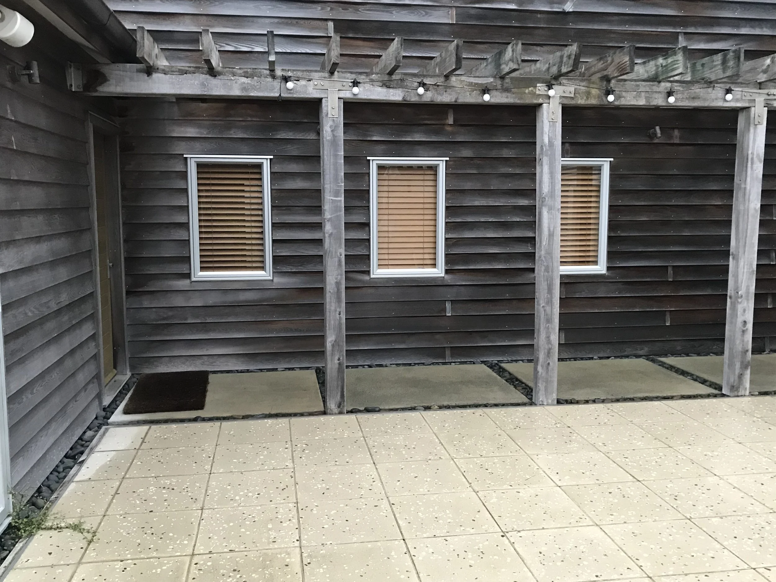 The image shows the exterior of a house with dark wooden siding, two windows with closed wooden blinds, and a partially constructed wooden patio or deck with vertical supports. The patio has string lights hanging underneath the top beam. The ground b