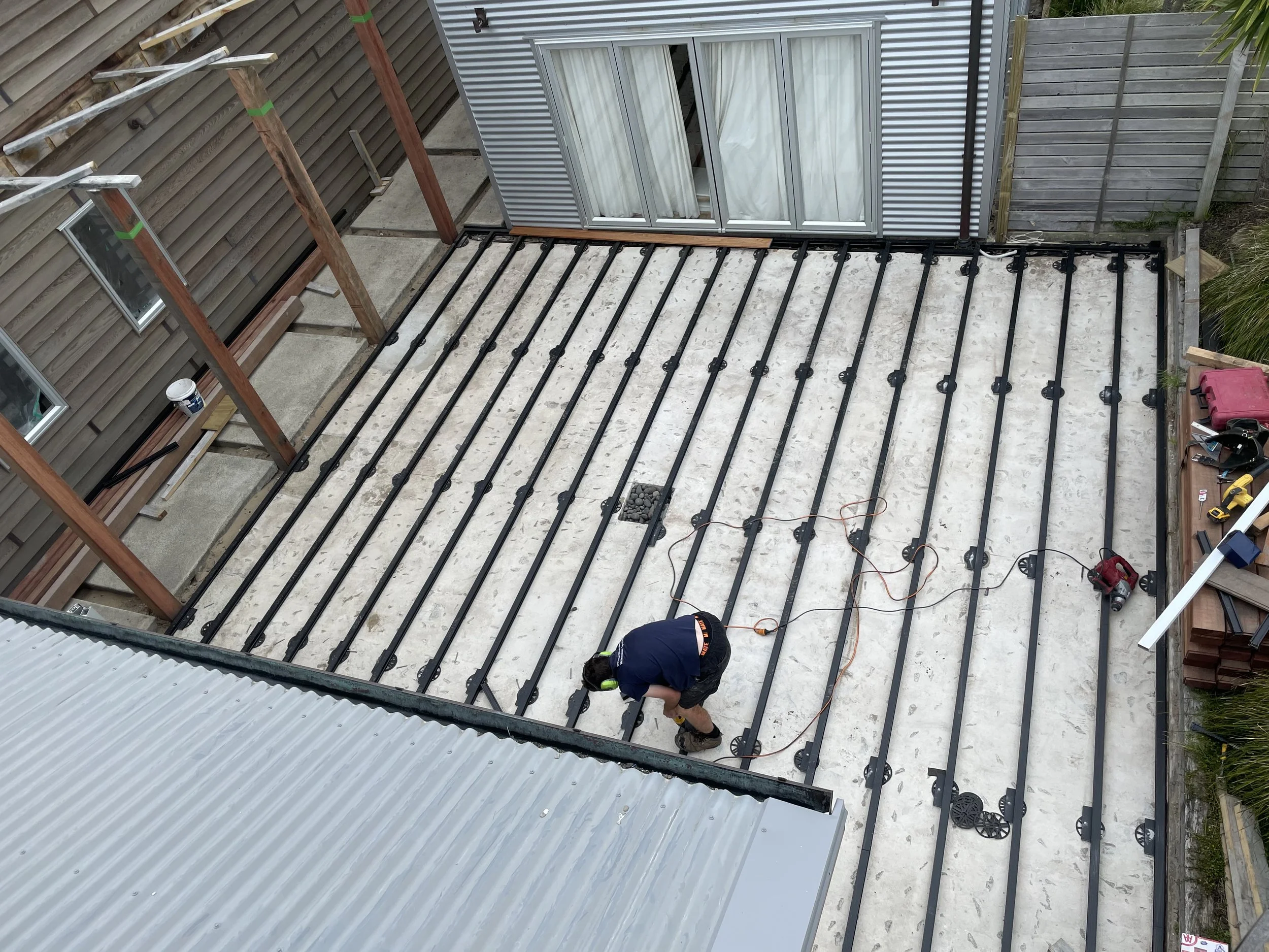 A construction worker is installing underfloor heating pipes on a concrete slab in a backyard patio area, with wooden and metal fencing surrounding the space.