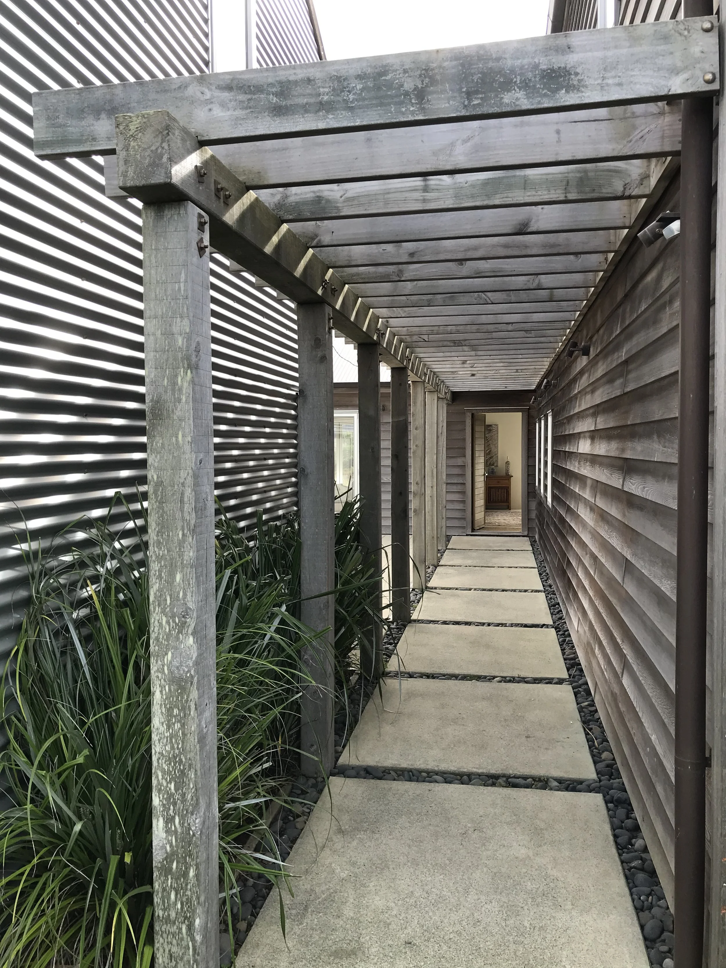 Concrete pathway with square slabs along a house wall, with a wooden pergola structure on the left, and a louvered fence on the back left side.