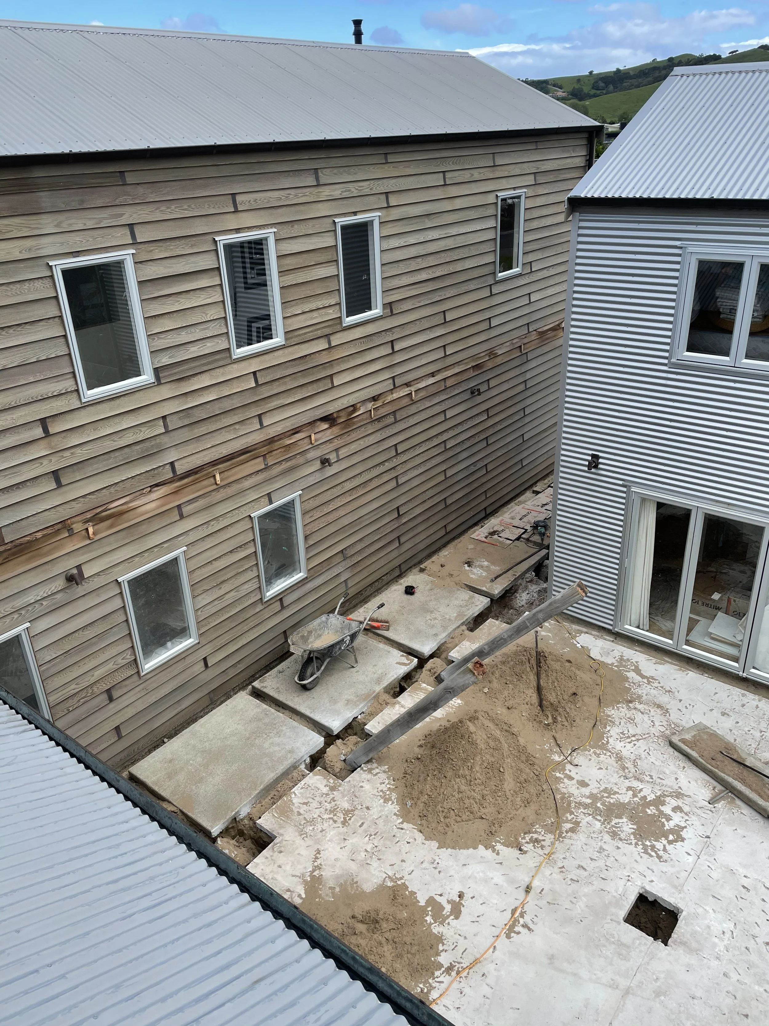 Construction site with an earthquake damage or repair in progress, showing exposed concrete foundation and scattered construction tools, between two buildings with metal and wooden siding, hills in the background.