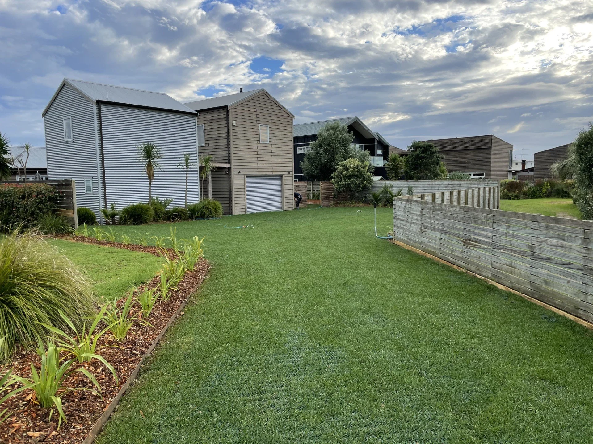 A backyard with a well-maintained lawn, small plants along the border, a wooden privacy fence, and modern houses with different siding styles and colors under a partly cloudy sky.