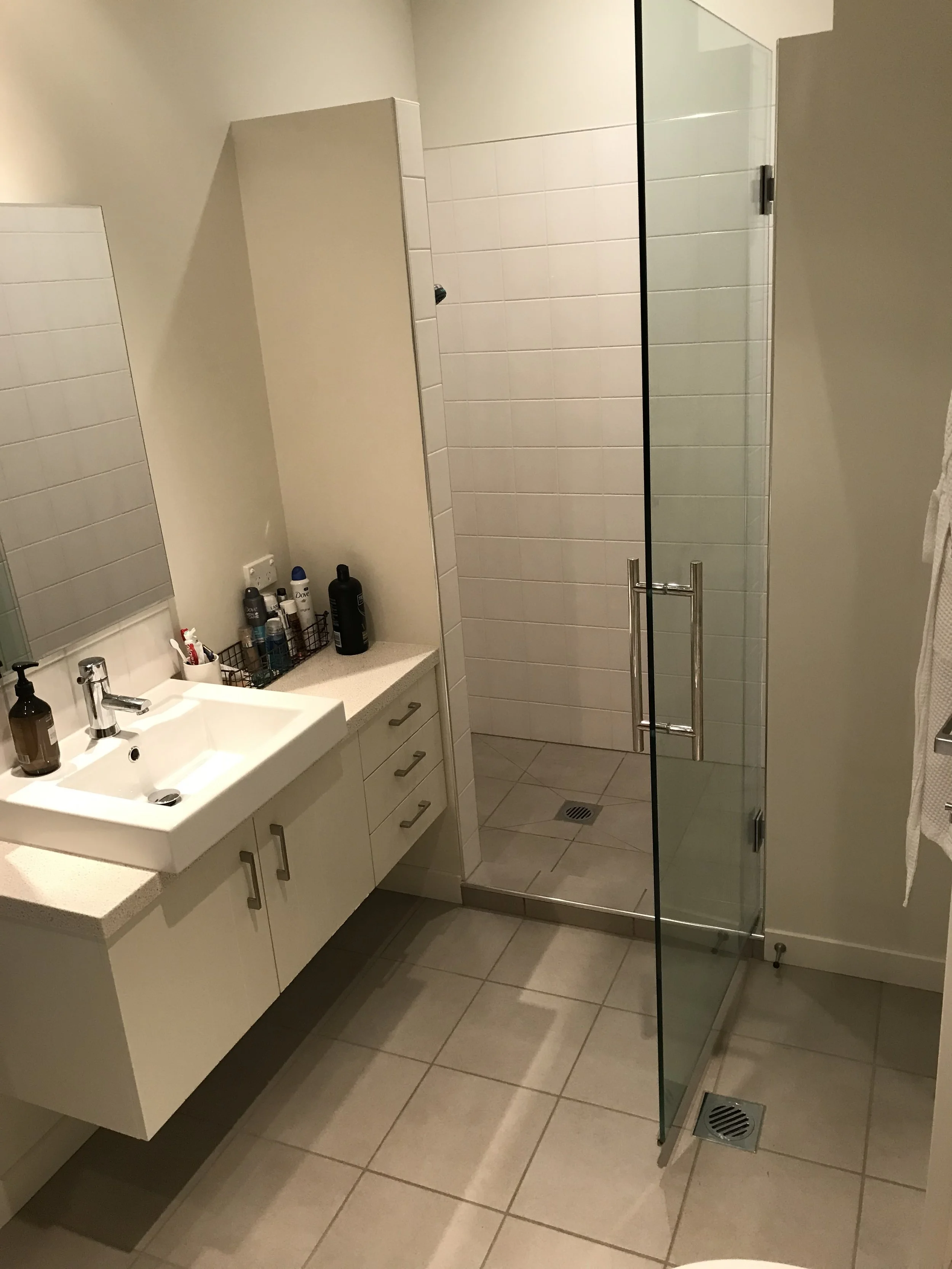 A small bathroom with a white countertop vanity, a mirror, and a glass enclosed shower with a metal handle, beige tiled floor, and various toiletries near the sink.