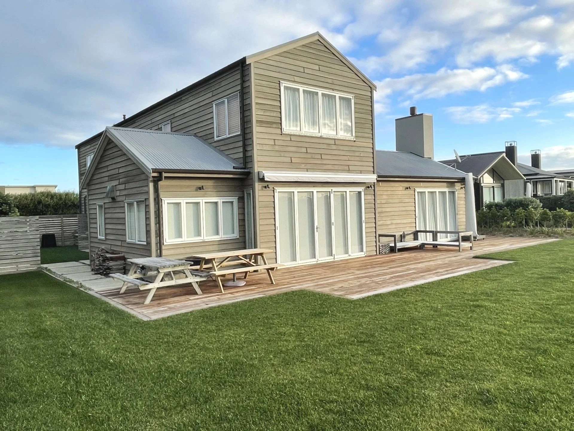 A beige two-story house with large windows, a sloped metal roof, and a spacious back deck is surrounded by a lawn and a wooden fence. There are outdoor tables and benches on the deck, under a cloudy sky.