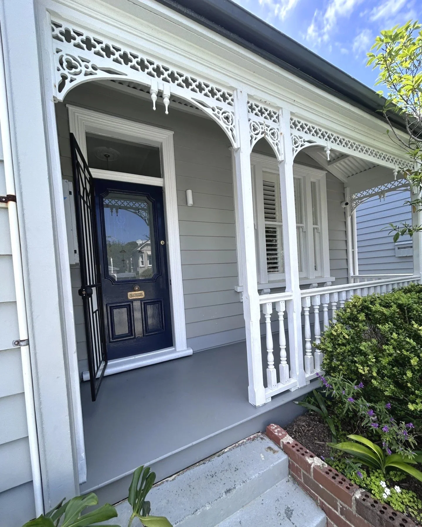 Front porch of a house with a dark blue door, a security gate, decorative white trim, and a white railing. There are plants and flowers in the garden bed next to the porch.