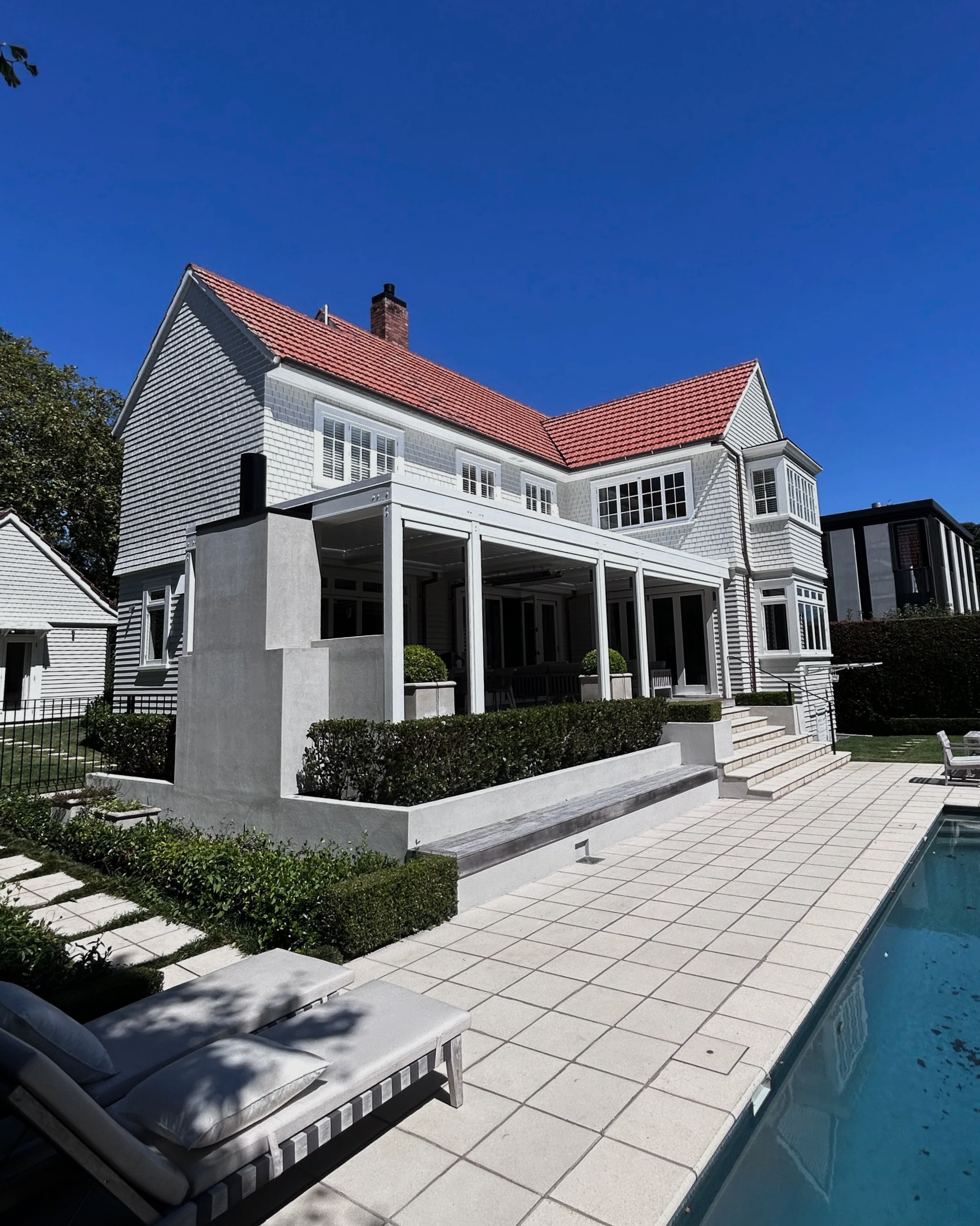 A modern house with white exterior and red tiled roof, surrounded by a backyard with a swimming pool, poolside lounge chairs, and landscaped garden with shrubs and concrete pathways.