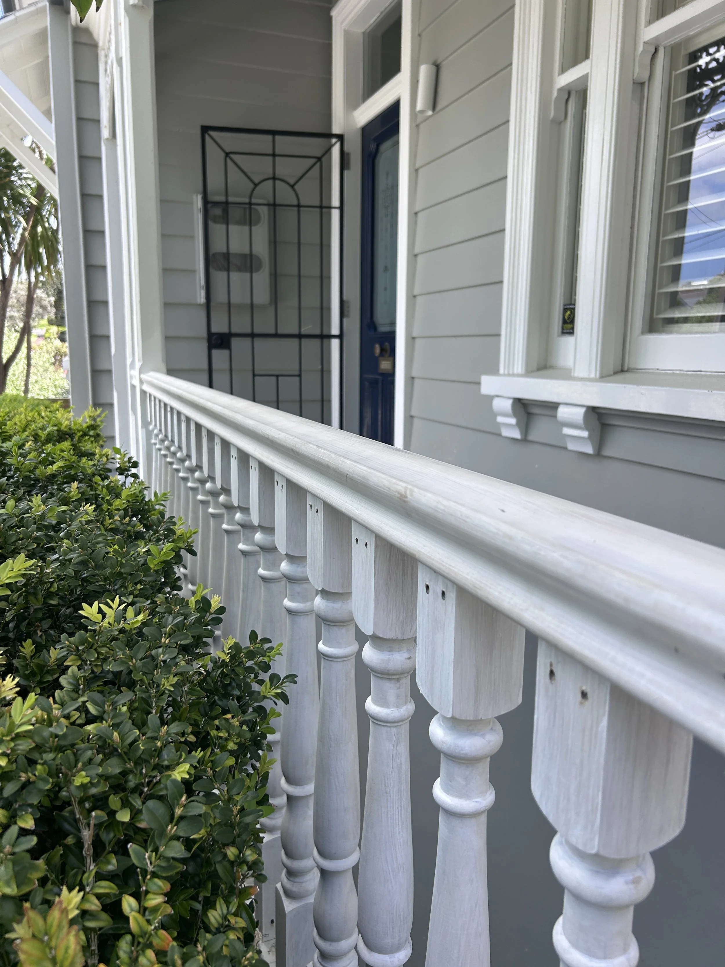Front porch with white railing, gray exterior wall, blue front door, window with white shutters, and a black metal security door with decorative arch design. Green bushes in the foreground.