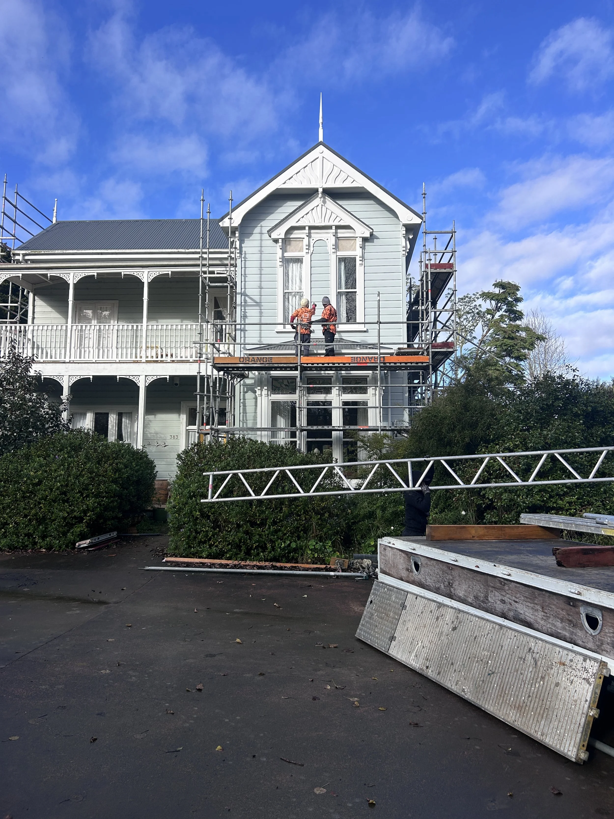 Construction workers on scaffolding in front of a Victorian-style house with blue exterior, white trim, and a porch. The sky is blue with scattered clouds, and there are some trees and bushes around the house. Construction materials are on the ground.