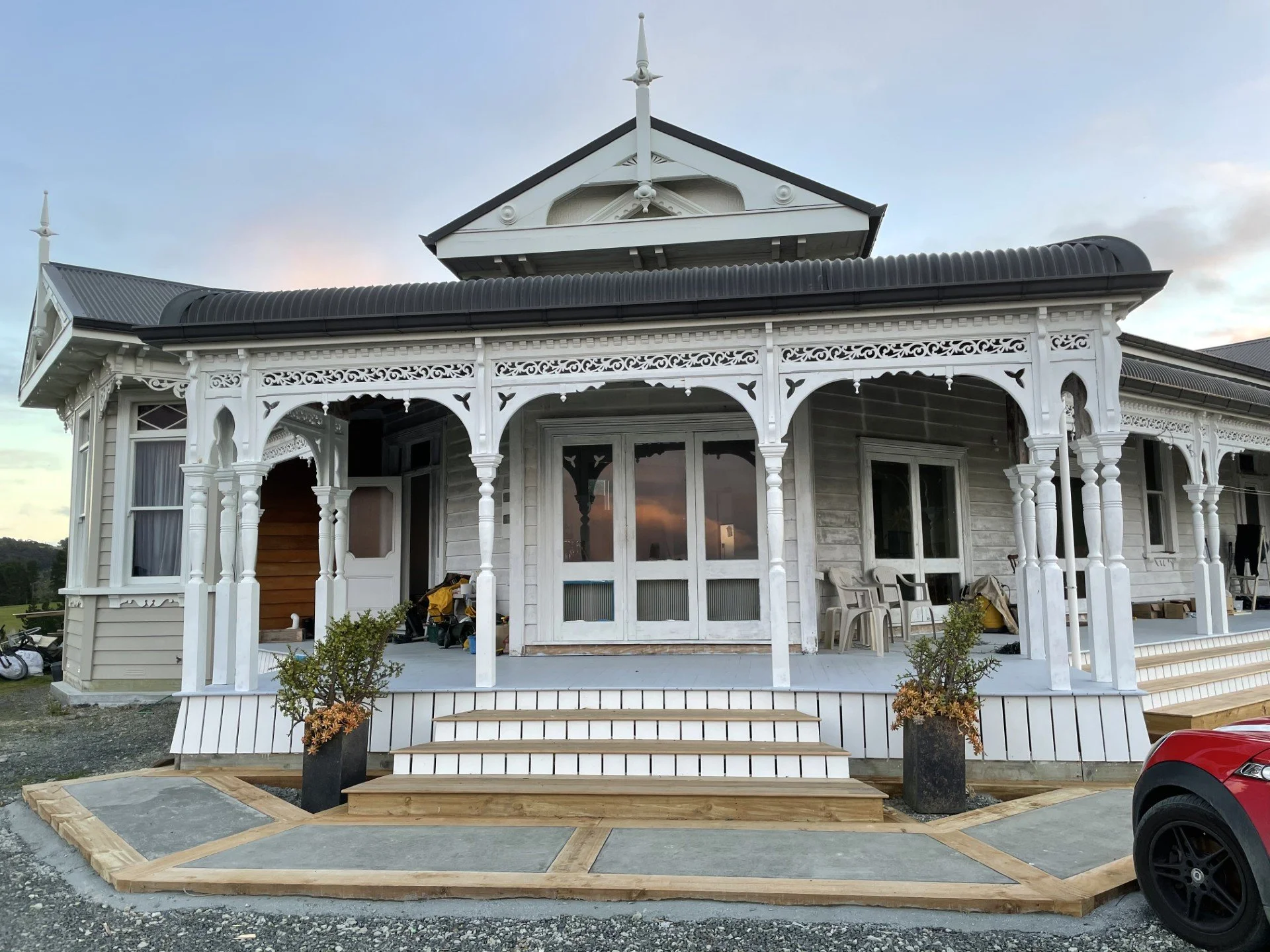 Front view of a partially constructed or renovated Victorian-style house with detailed white woodwork, lace trim, and a porch, with stairs and potted plants in front.