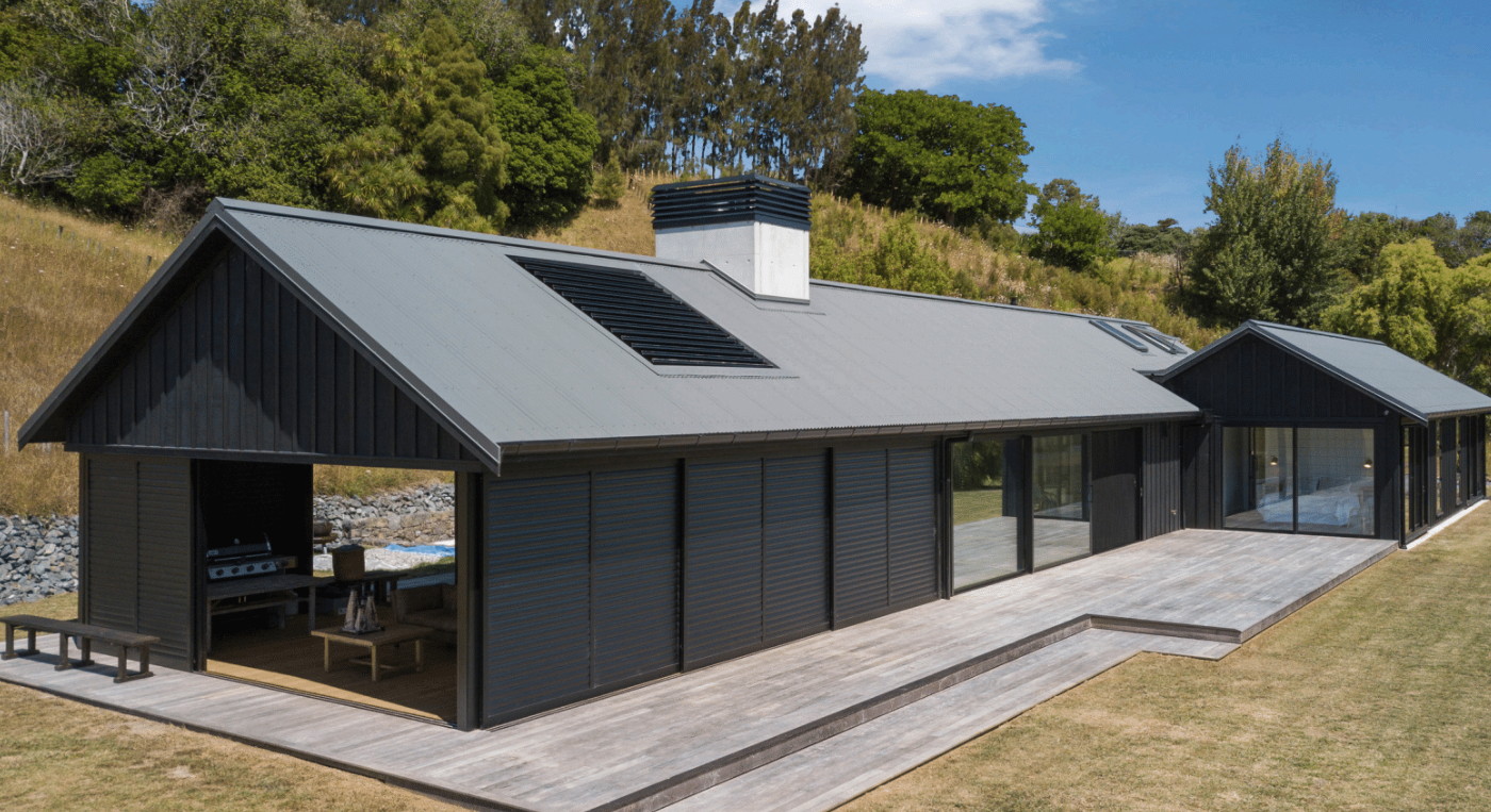 Modern black house with large sliding glass doors opening to a wooden deck, surrounded by grass and trees on a sunny day.