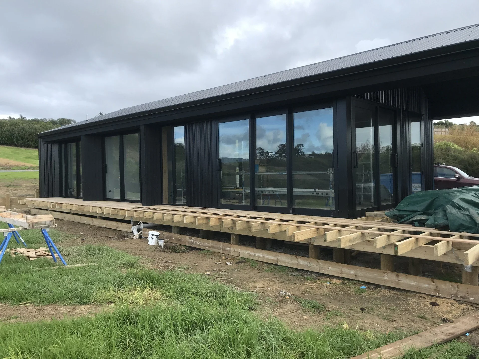 Construction site with a modern black building with glass doors and windows, partially built on a wooden platform, with construction tools and materials nearby, and cloudy sky overhead.