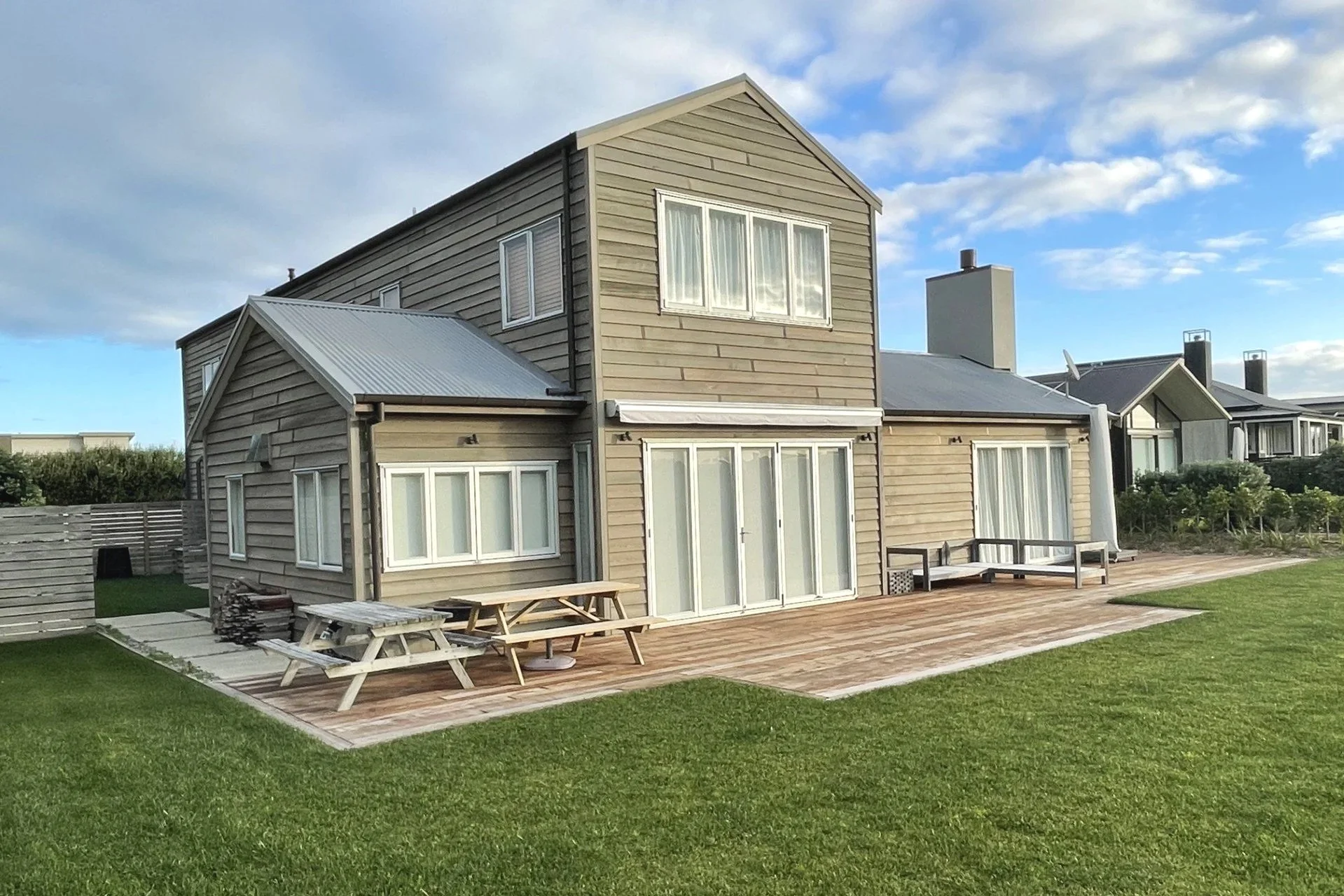 A modern two-story house with beige siding and white trim, situated in a backyard with a wooden patio, grass lawn, and a wooden fence. The house has multiple windows, including a large one on the second floor. The sky is partly cloudy.