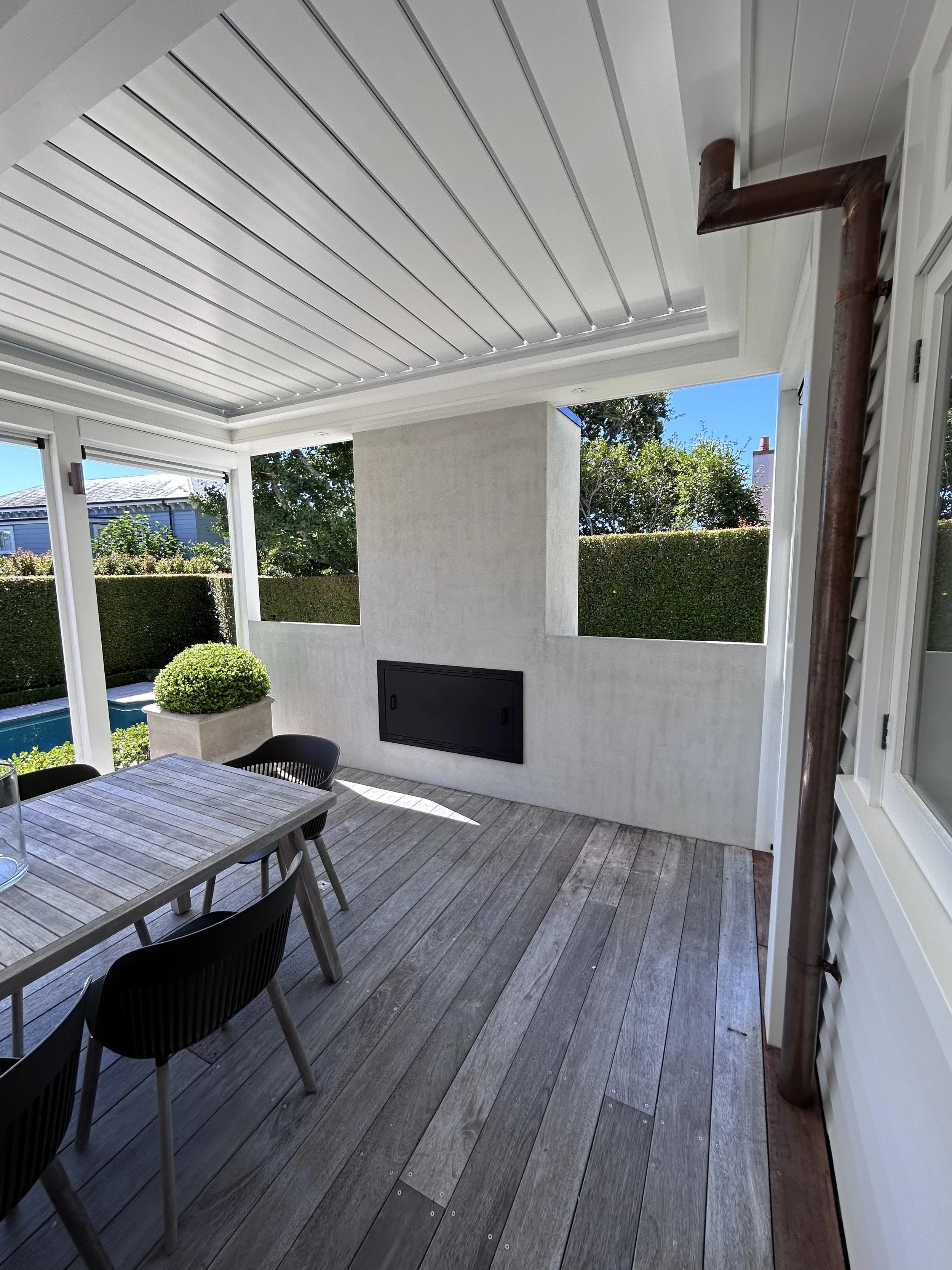 Covered outdoor patio with wooden flooring, a rectangular wooden table, black chairs, and a potted plant near a white wall with a fireplace. The ceiling has white siding, and a rusty downspout runs along the side.