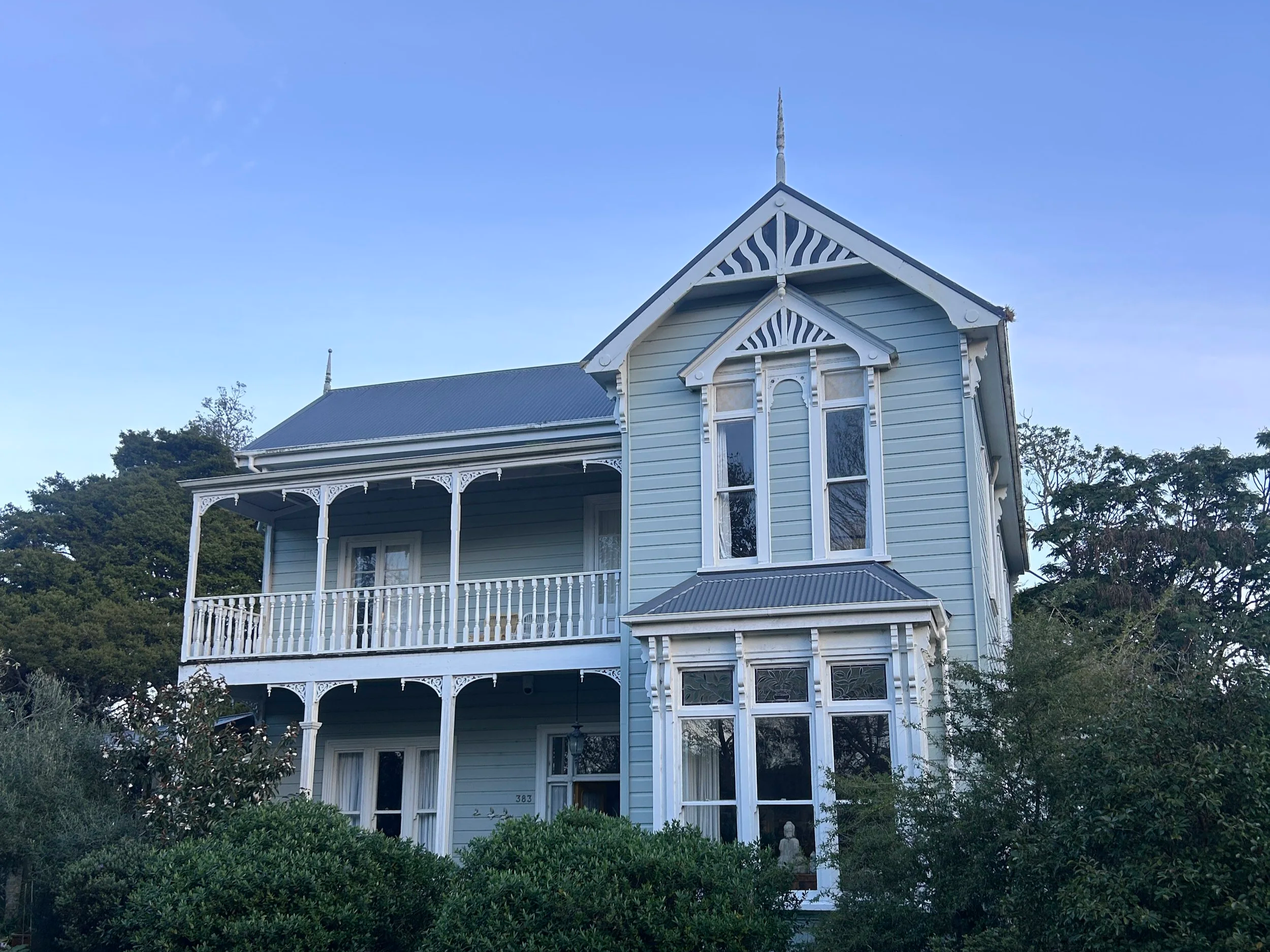 A three-story light blue Victorian-style house with white trim, detailed woodwork, and a prominent front porch surrounded by greenery and trees.