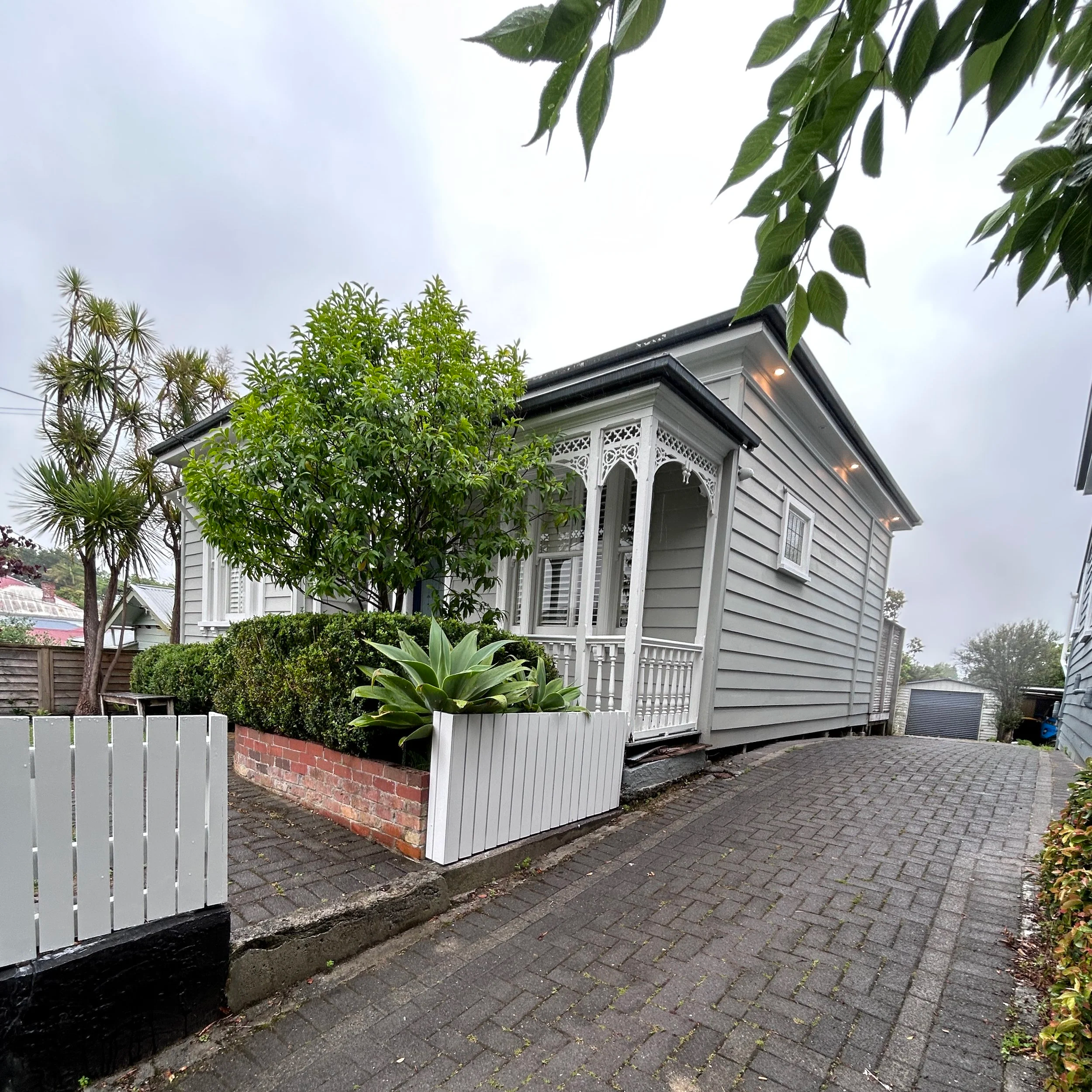 A house with a decorative porch, surrounded by trees and plants, with a paved walkway and a white picket fence.