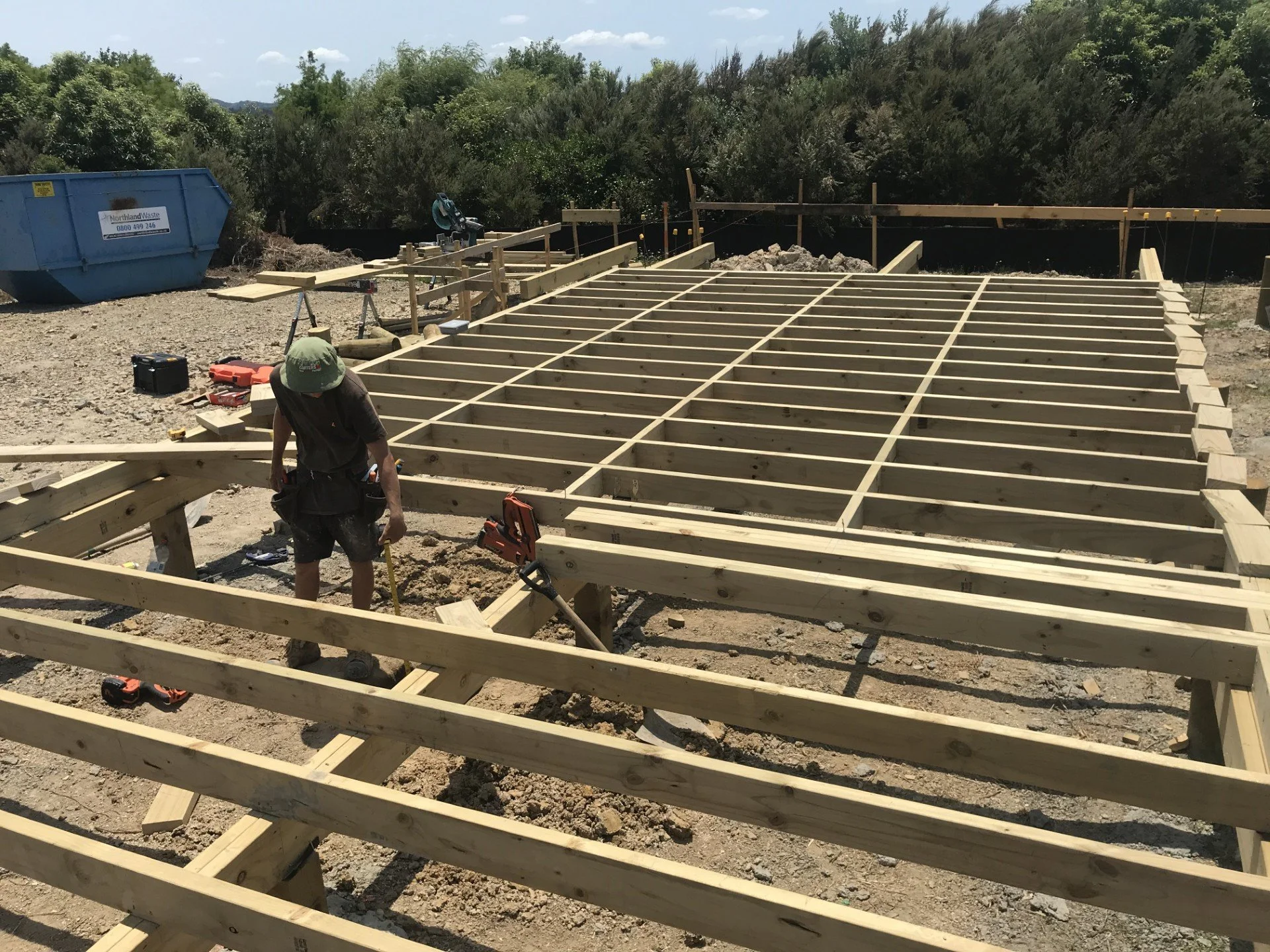 Construction site with wooden framework for a building, a worker wearing a green hat and black shirt, tools scattered on the ground, and trees in the background.
