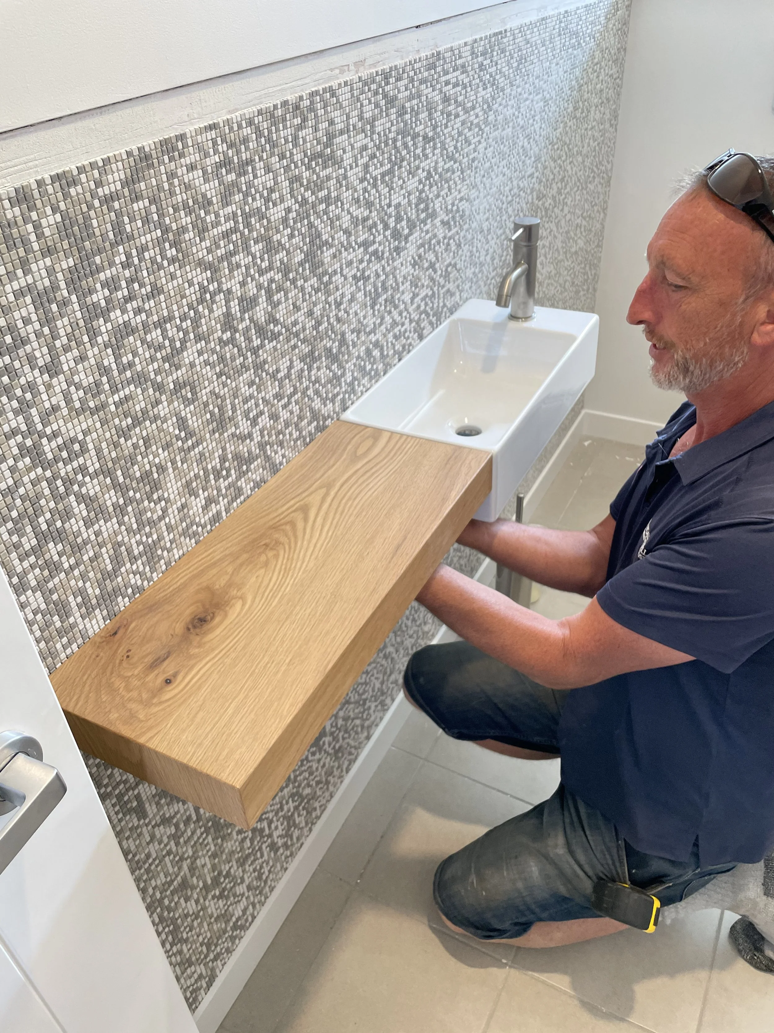 A man kneeling at a sink in a bathroom, examining the faucet with a wooden countertop, mosaic tile backsplash, and tiled floor.