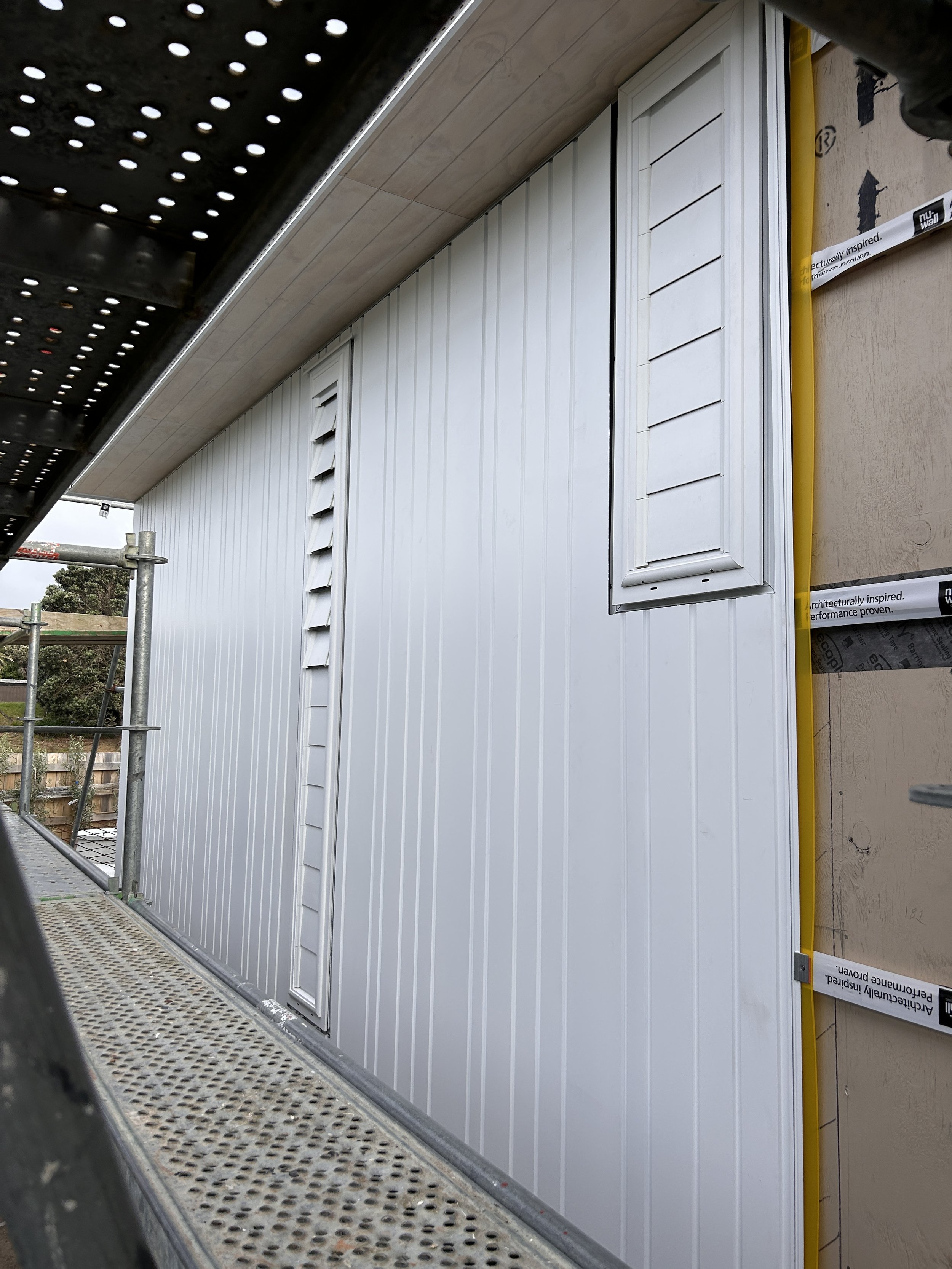 Close-up of a construction site exterior showing white siding panels and a window with shutters. Part of a metal scaffolding platform is visible in the foreground.