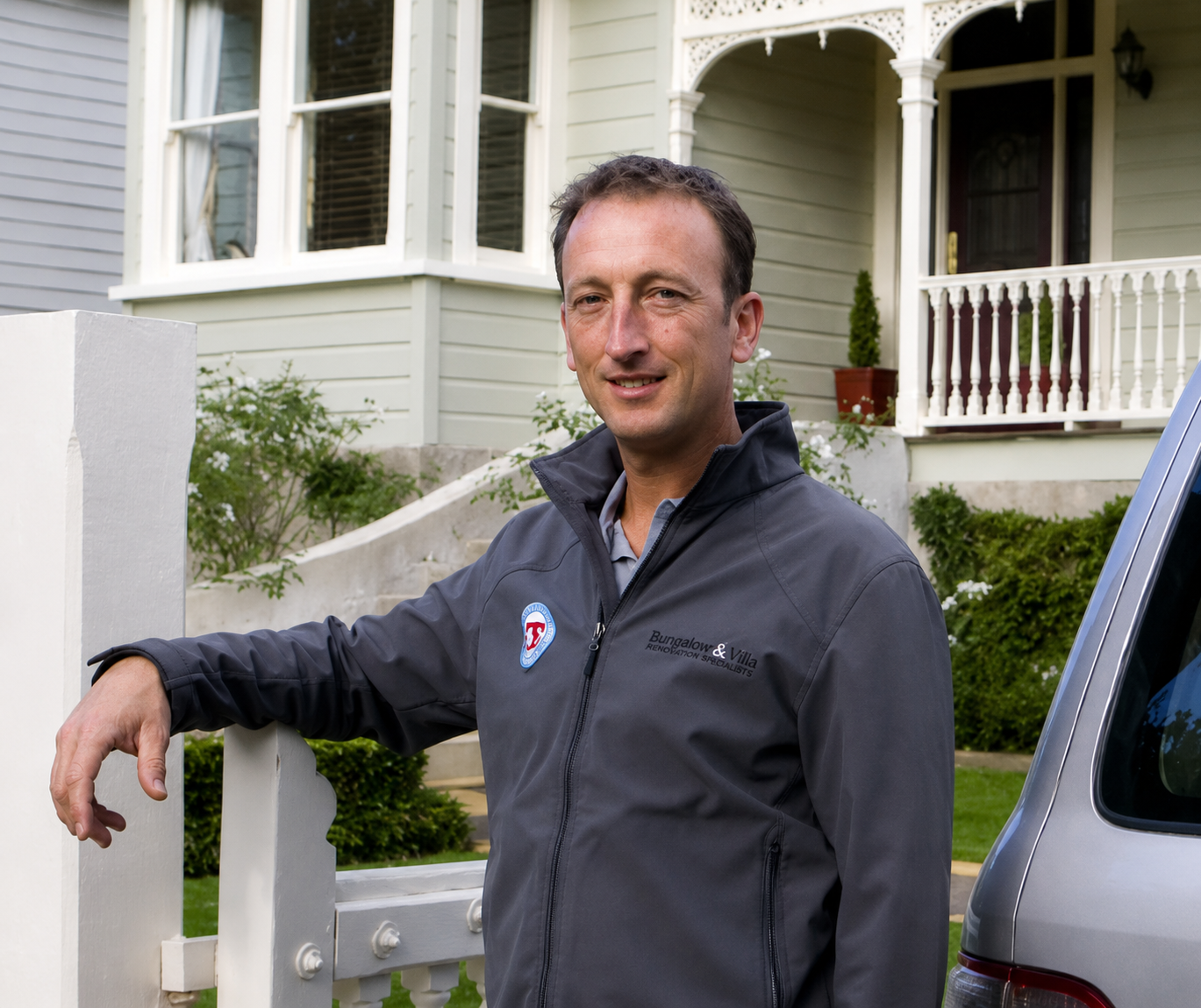 A man standing outside in front of a house, leaning on a white wooden fence, wearing a gray jacket with a logo that reads 'Bungalow & Villa Renovation Specialists'