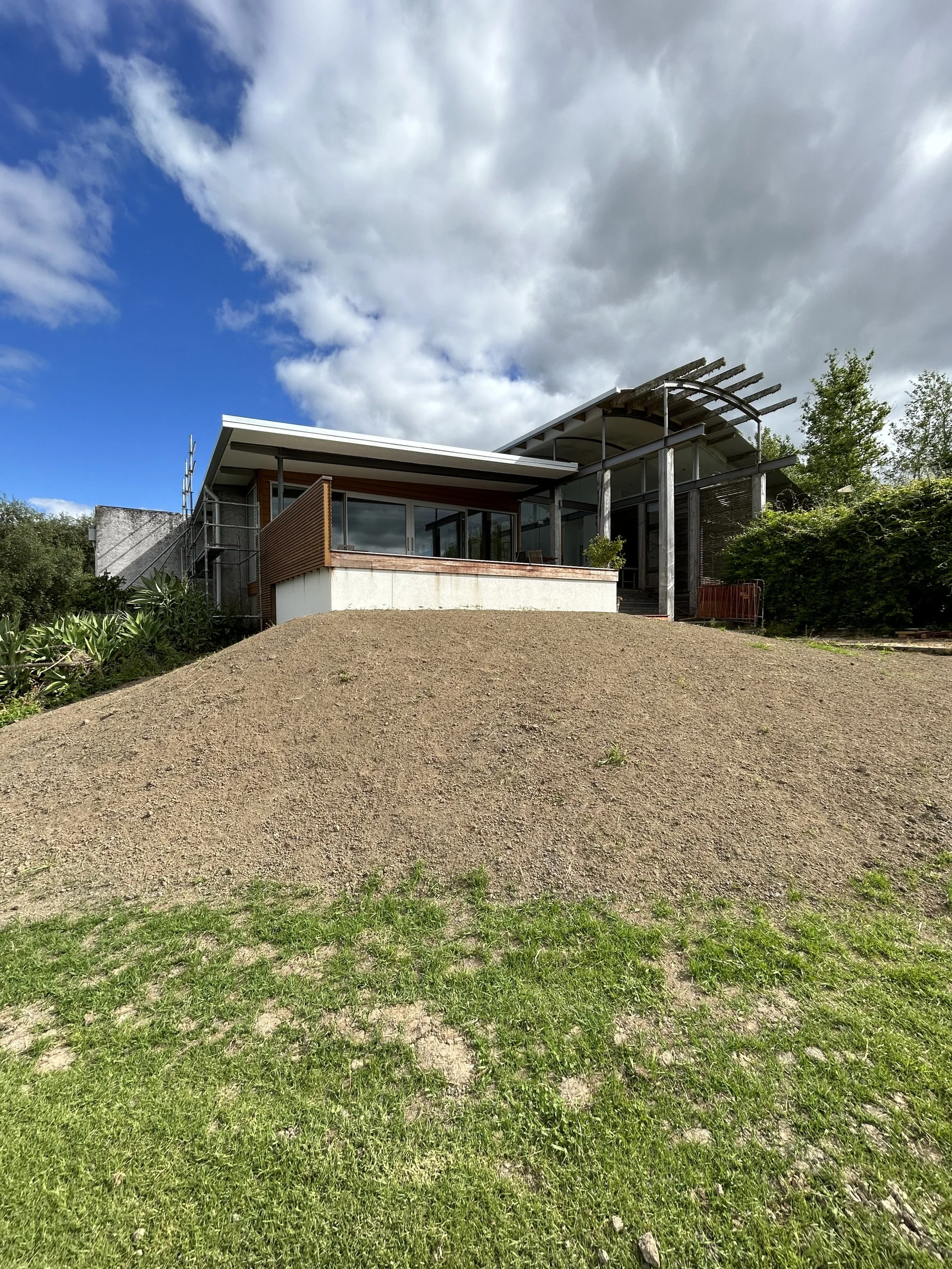 Modern house with large glass windows and wooden accents, situated on a small hill, with a partly cloudy sky overhead and green grass in the foreground.