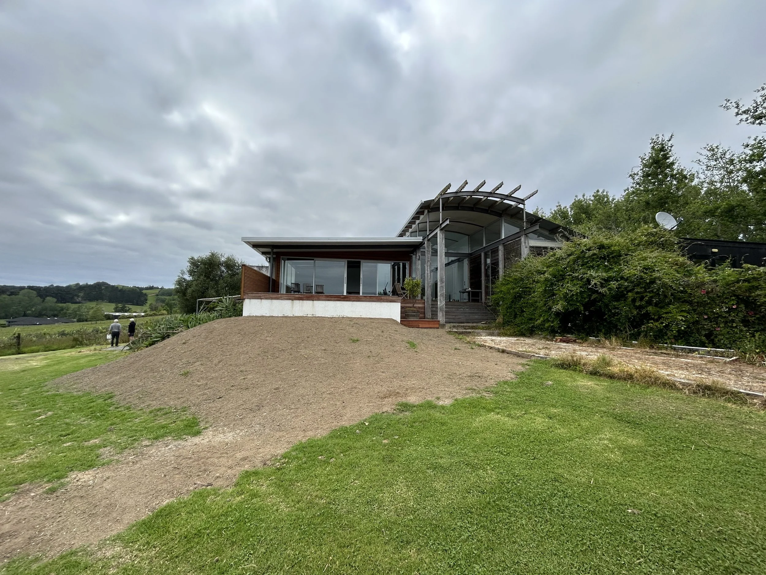 A modern house with large glass windows, a curved roof structure, surrounded by greenery and trees under a cloudy sky, with a small dirt slope and two people walking on a path.