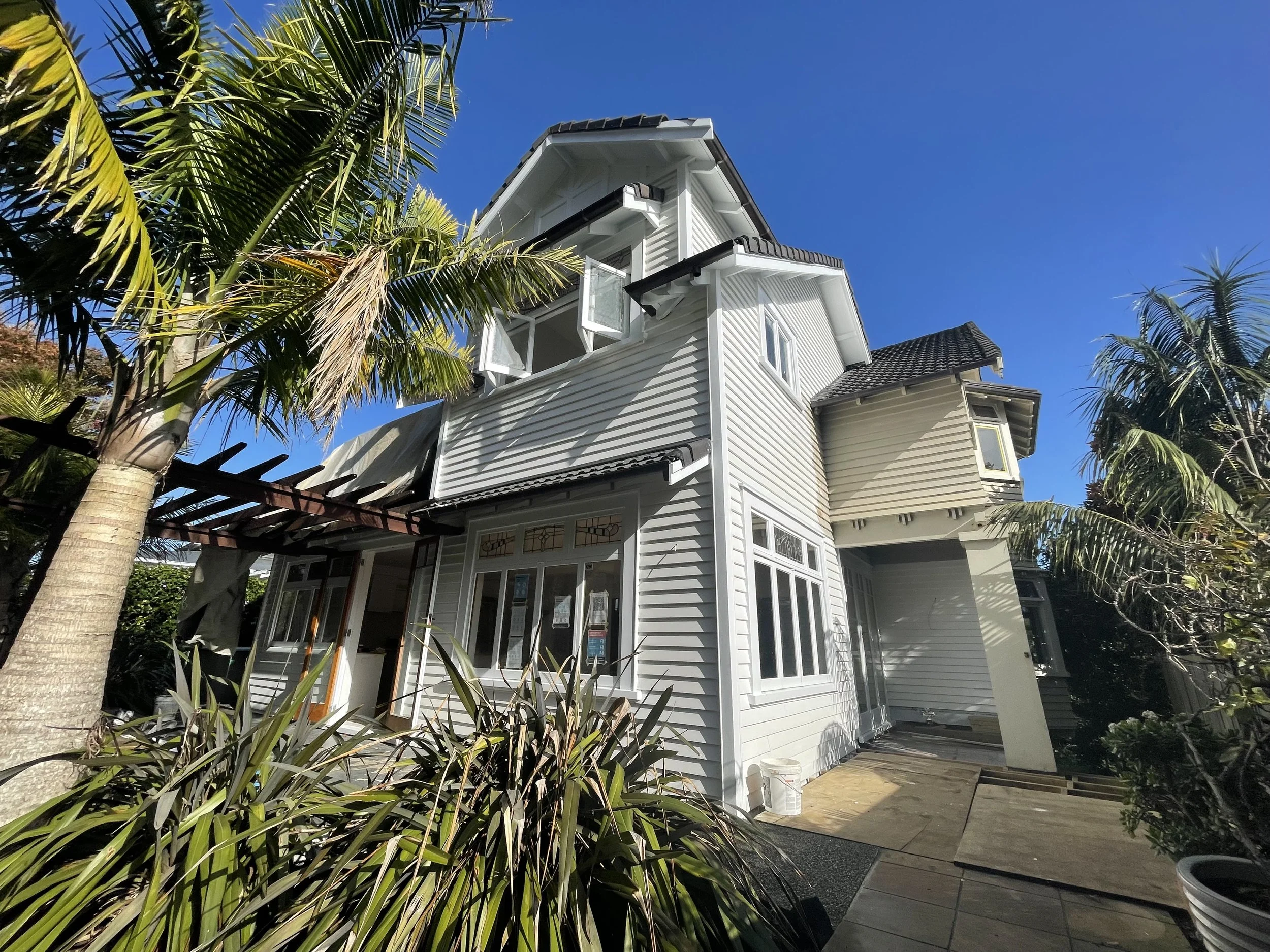 A white multi-story house with black roof tiles, surrounded by tropical plants and trees, with a blue sky in the background.