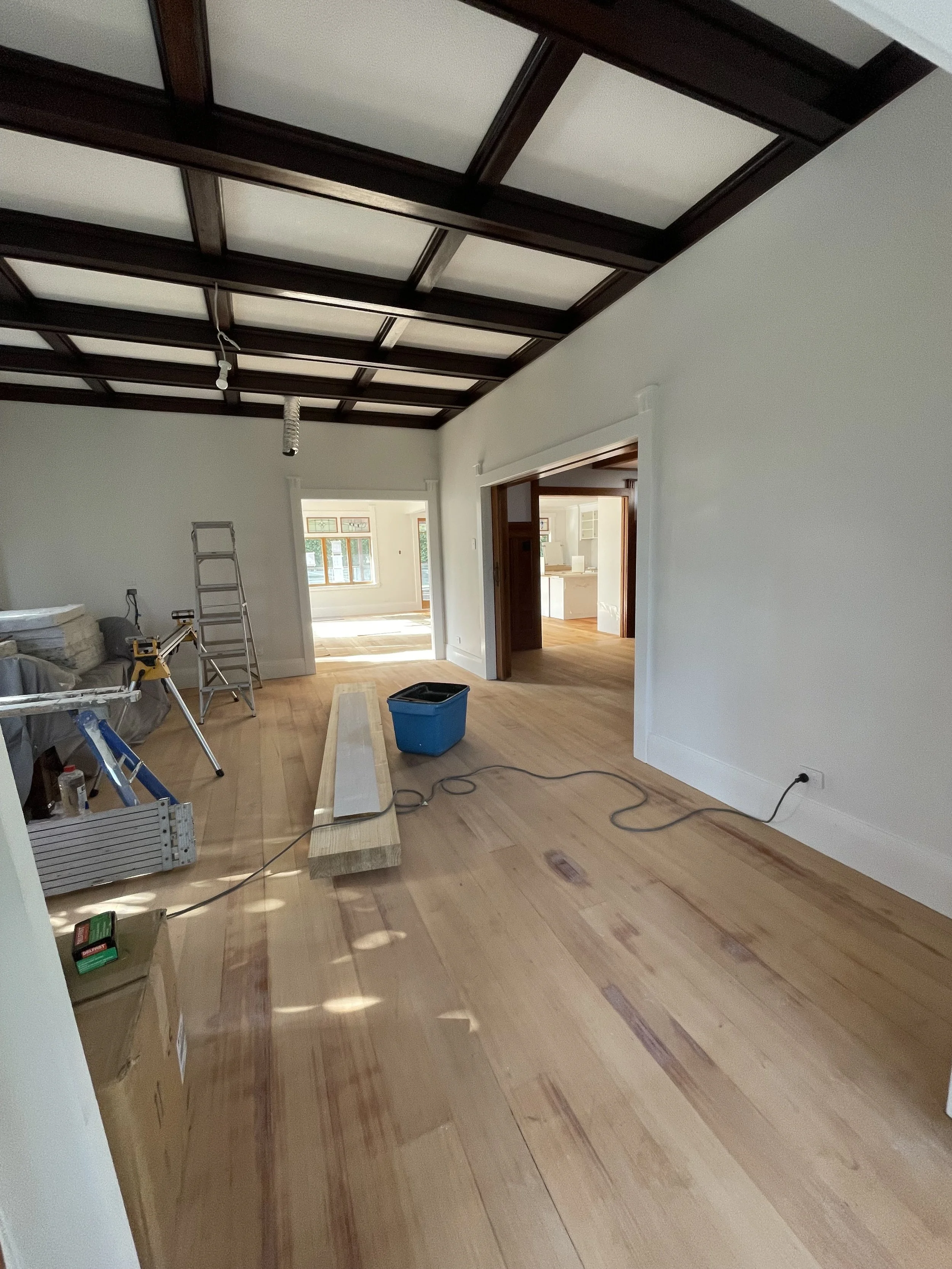 Room under renovation with unfinished flooring, construction tools, ladder, and construction materials. Walls are painted white, and there's a doorway leading to another room with natural light.