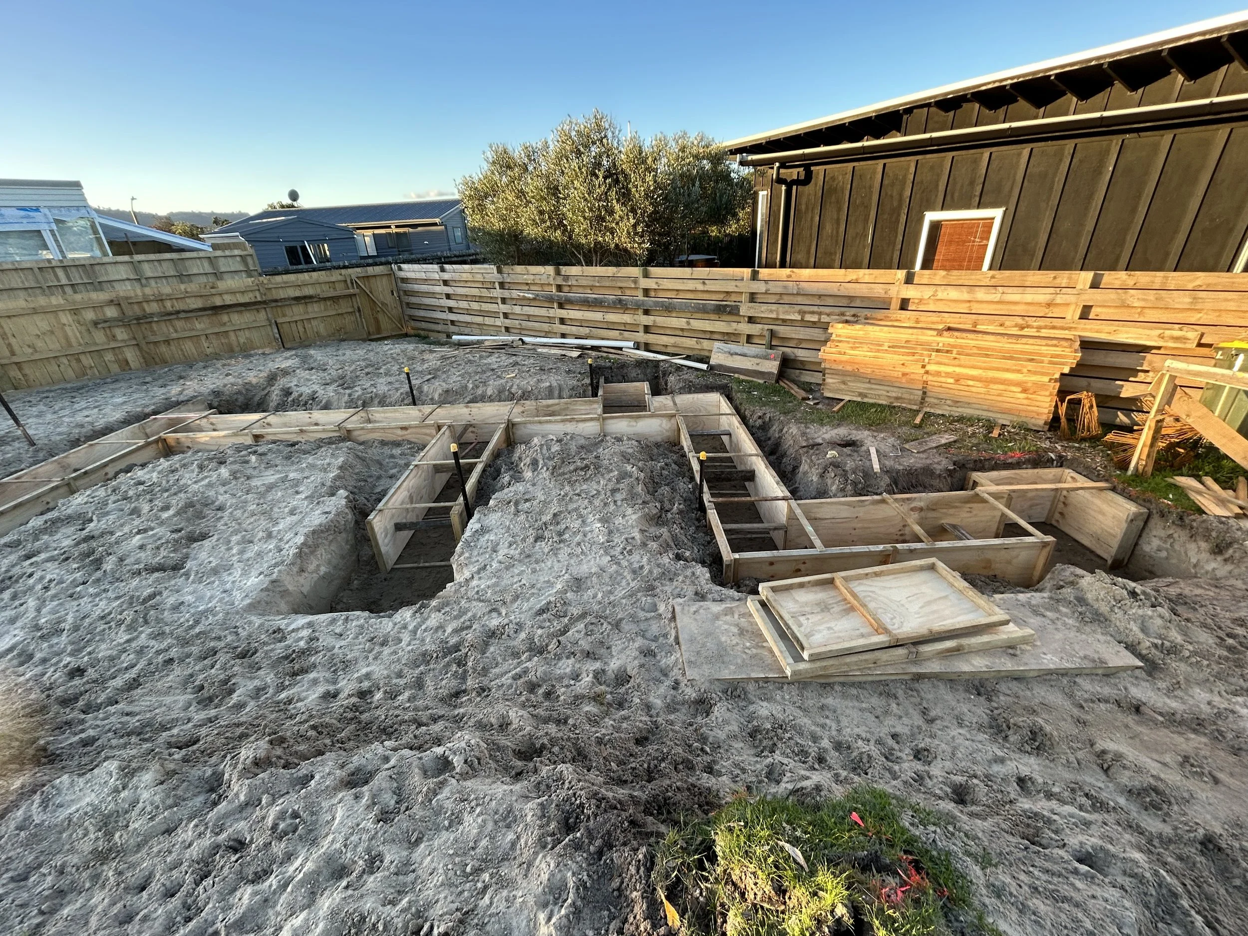 Construction site with a dug-out area and wooden forms for pouring concrete, surrounded by wooden fencing and neighboring houses under a clear sky.