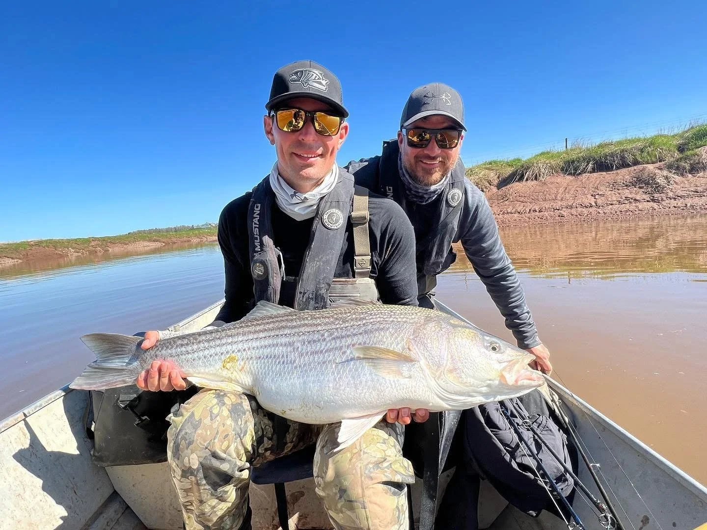 Two men in a boat holding a large fish with a river and blue sky in the background.