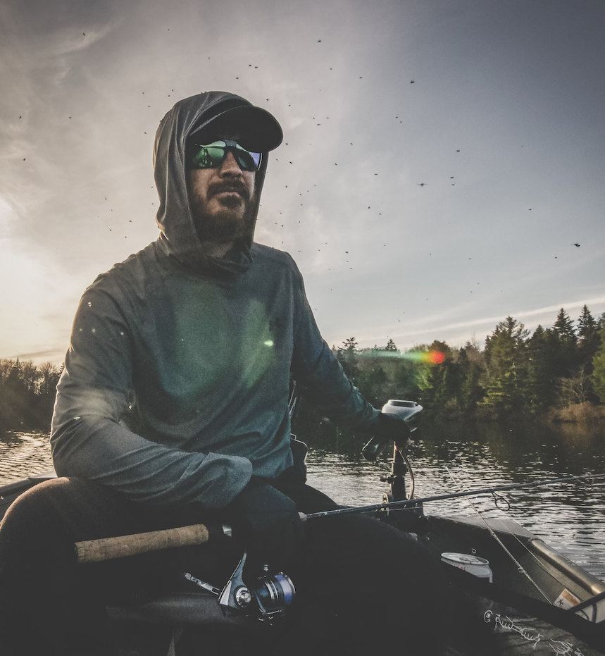 Man fishing in a boat on a river during sunset or dusk, wearing sunglasses, a hoodie, and a jacket.