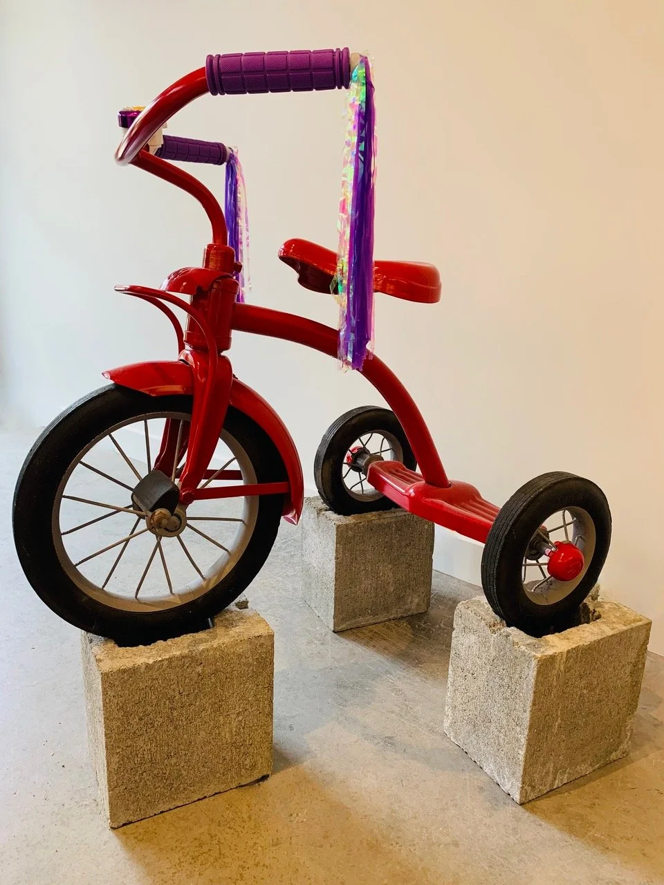 Mixed media art sculpture of a red tricycle with a purple handlebar and a purple streamers on the handlebar, mounted on concrete blocks against a white wall.