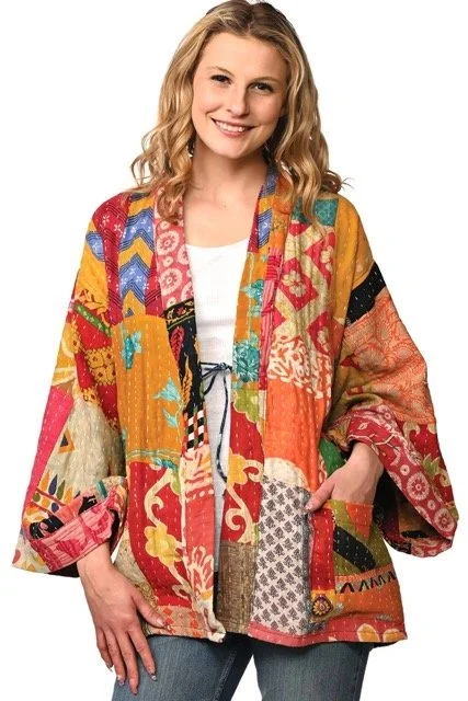 A woman wearing a colorful quilted patchwork jacket with various patterns and colors, smiling with her right hand on her hip.
