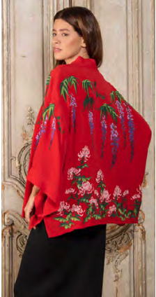 A woman with long dark hair wearing a red embroidered kimono-style garment with green, white, and purple floral and tree branch designs, standing against a vintage wooden paneled background.