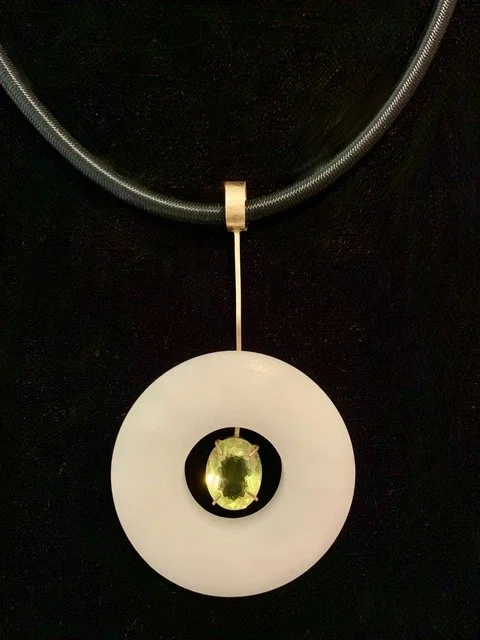 Pendant necklace with a large white circular pendant containing a yellow gemstone in the center, hanging from a silver chain against a black background. Created by Theresa Killgore.