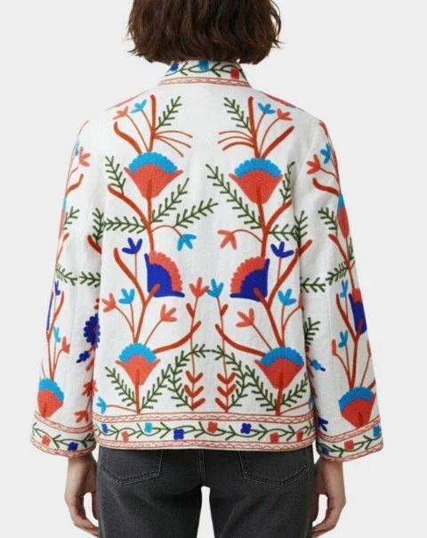 Back view of a person wearing a white jacket with bright floral embroidery in blue, orange, green, and red, featuring stylized flowers and leaves.