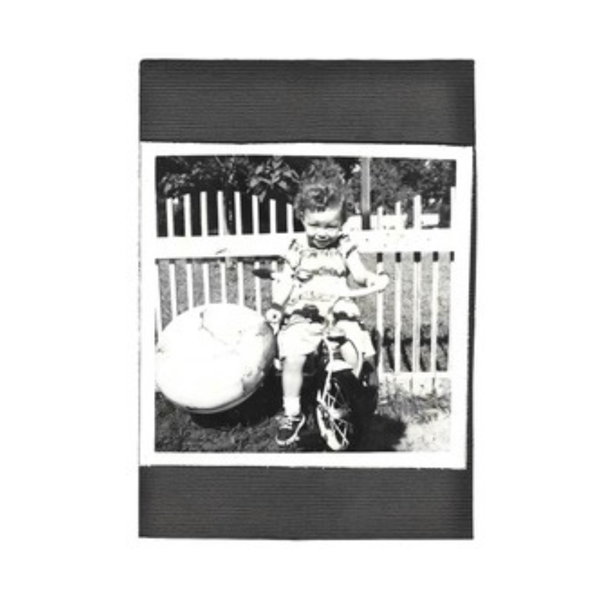 A young girl with curly hair sitting on a small bicycle, holding an ice cream cone, next to a large round object, in front of a wooden fence and a house with a dark roof.
