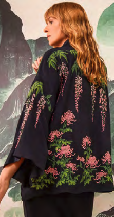 Woman with long wavy hair wearing a black jacket with pink flamingo and green foliage patterns, standing against a painted nature backdrop.