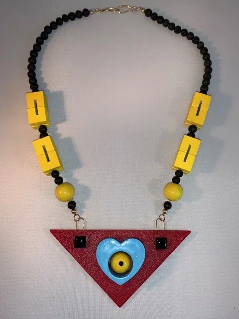 Colorful necklace with black beads, yellow blocks, and a red triangular pendant featuring a blue heart shape with a yellow center and black accents.