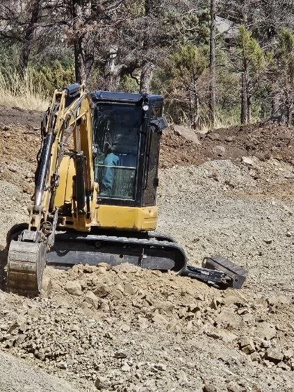 A yellow excavator working on rocky terrain at a construction site with trees in the background.
