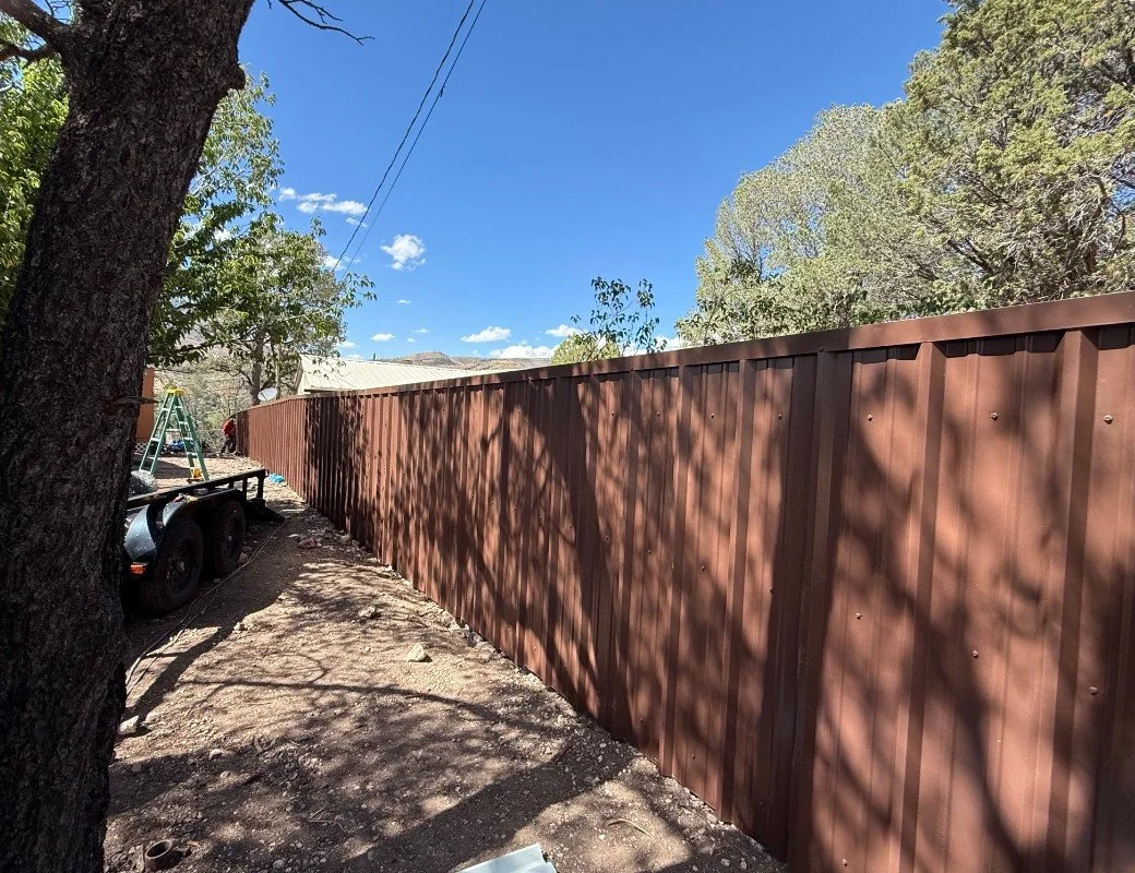 A long brown metal fence runs alongside a dirt yard with some tools and equipment, including a ladder and a trailer, under a clear blue sky with some clouds and trees casting shadows.
