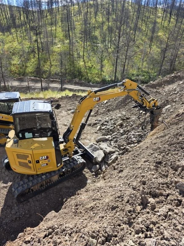 A yellow Caterpillar excavator digging into a hillside with trees in the background.