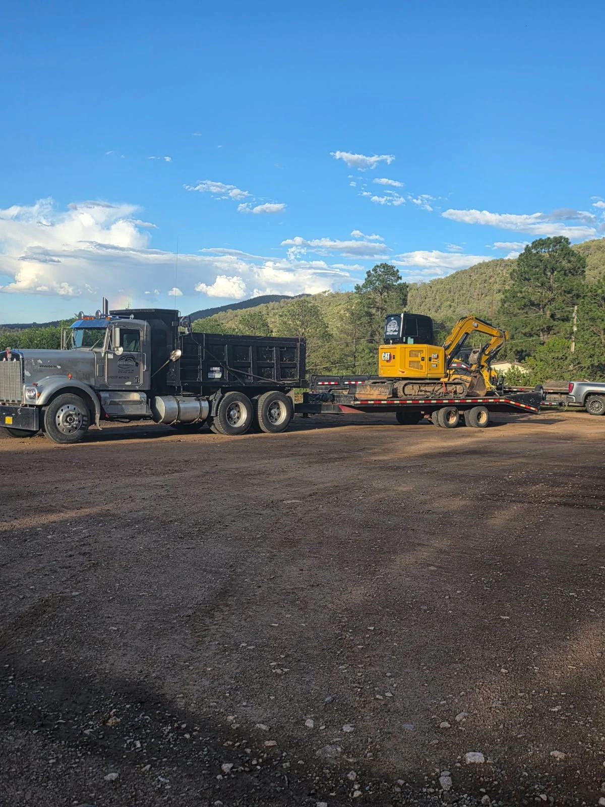 A black truck hauling a flatbed trailer with a yellow CAT excavator on it, parked on a dirt lot with mountains and trees in the background under a blue sky with scattered clouds.