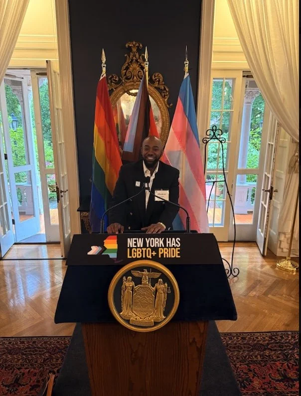 A man in a suit standing at a podium with the New York state seal, in front of multiple flags including the LGBTQ+ pride rainbow flag, the genderqueer flag, and the transgender flag, with a mirror and windows in the background.
