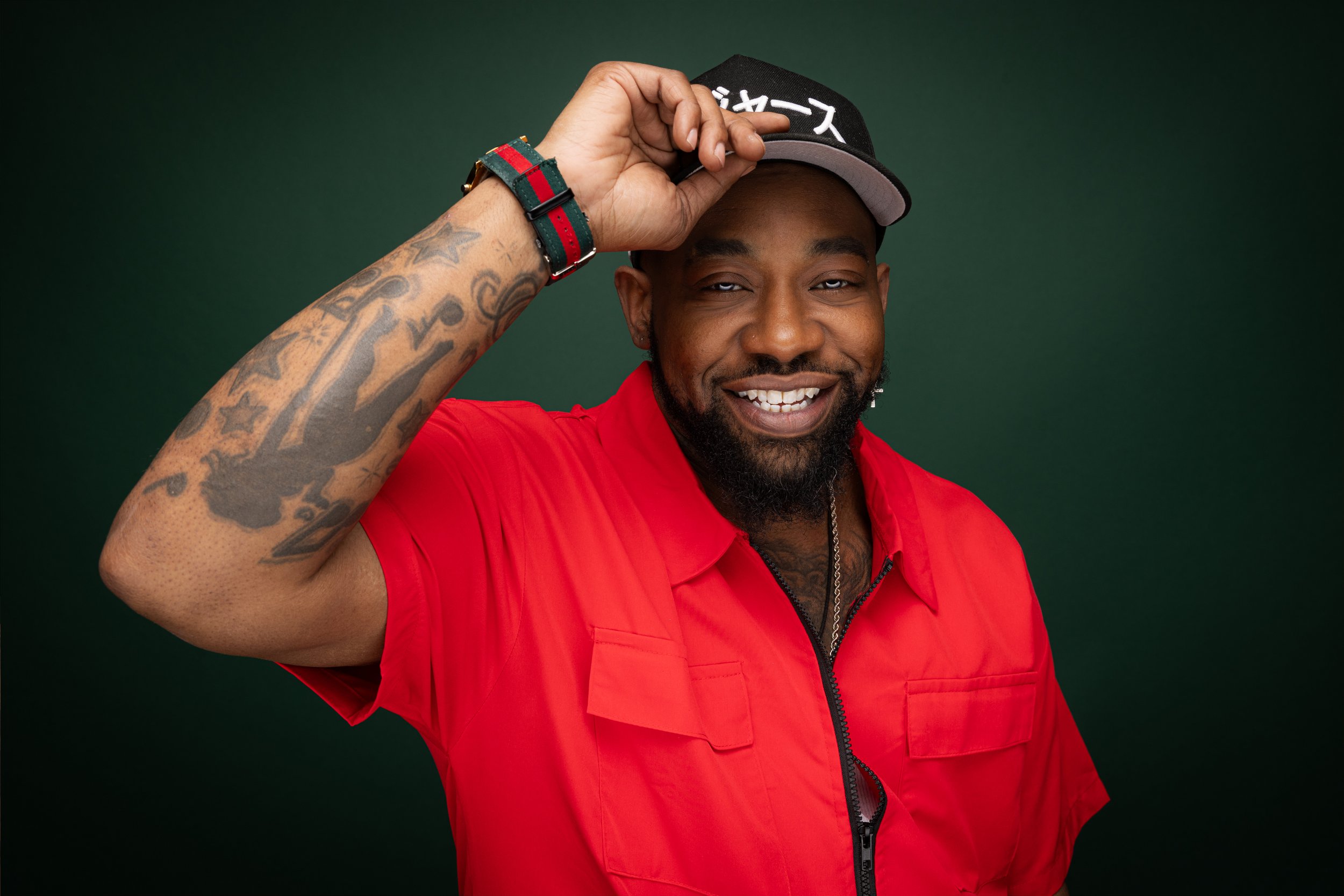 A smiling man in a red shirt and black cap partially adjusting the cap with his right hand, against a dark green background.