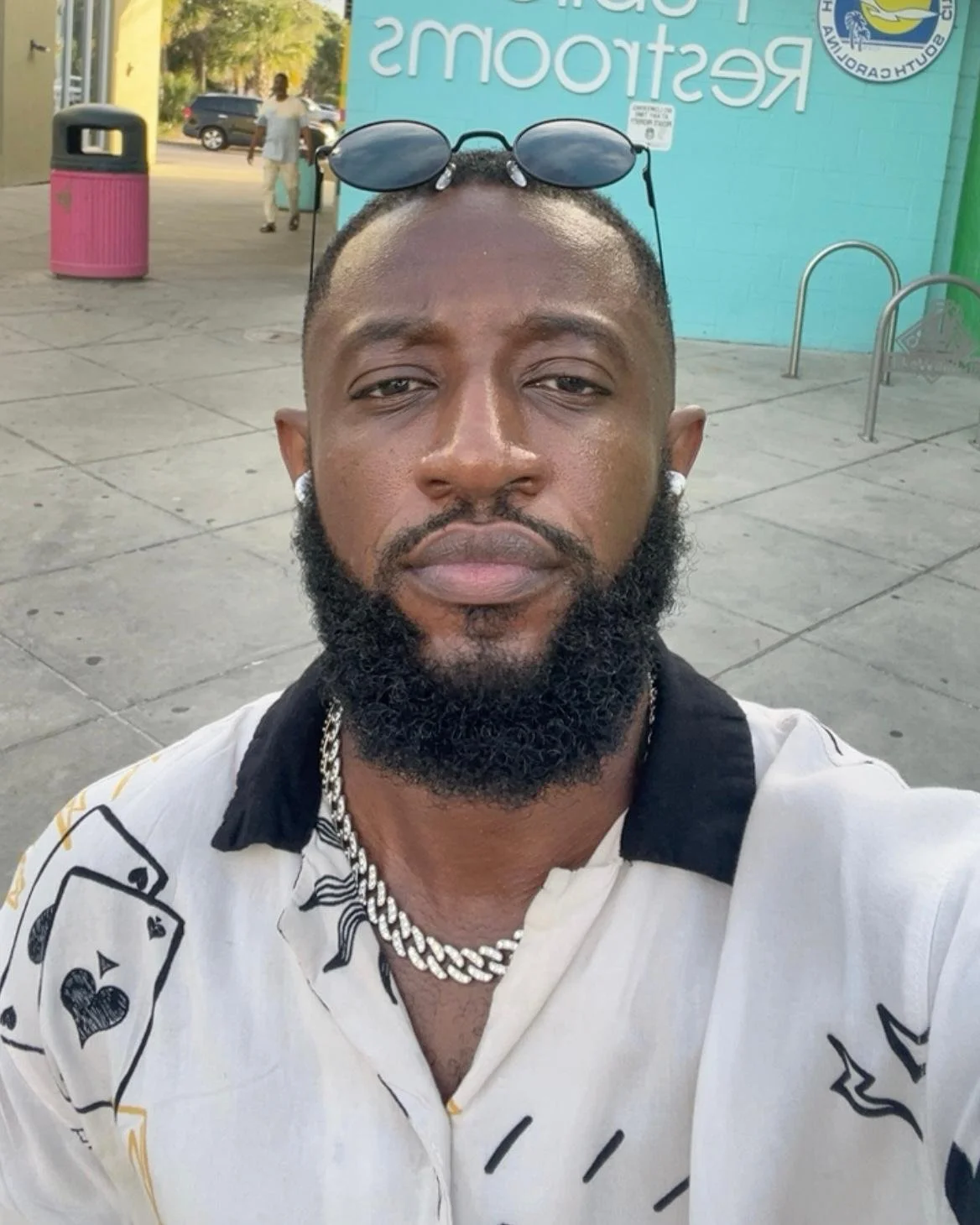 A man with a beard wearing sunglasses on his head, a white shirt with black and gold designs, a silver chain necklace, and earrings, taking a selfie outdoors with a street scene in the background.