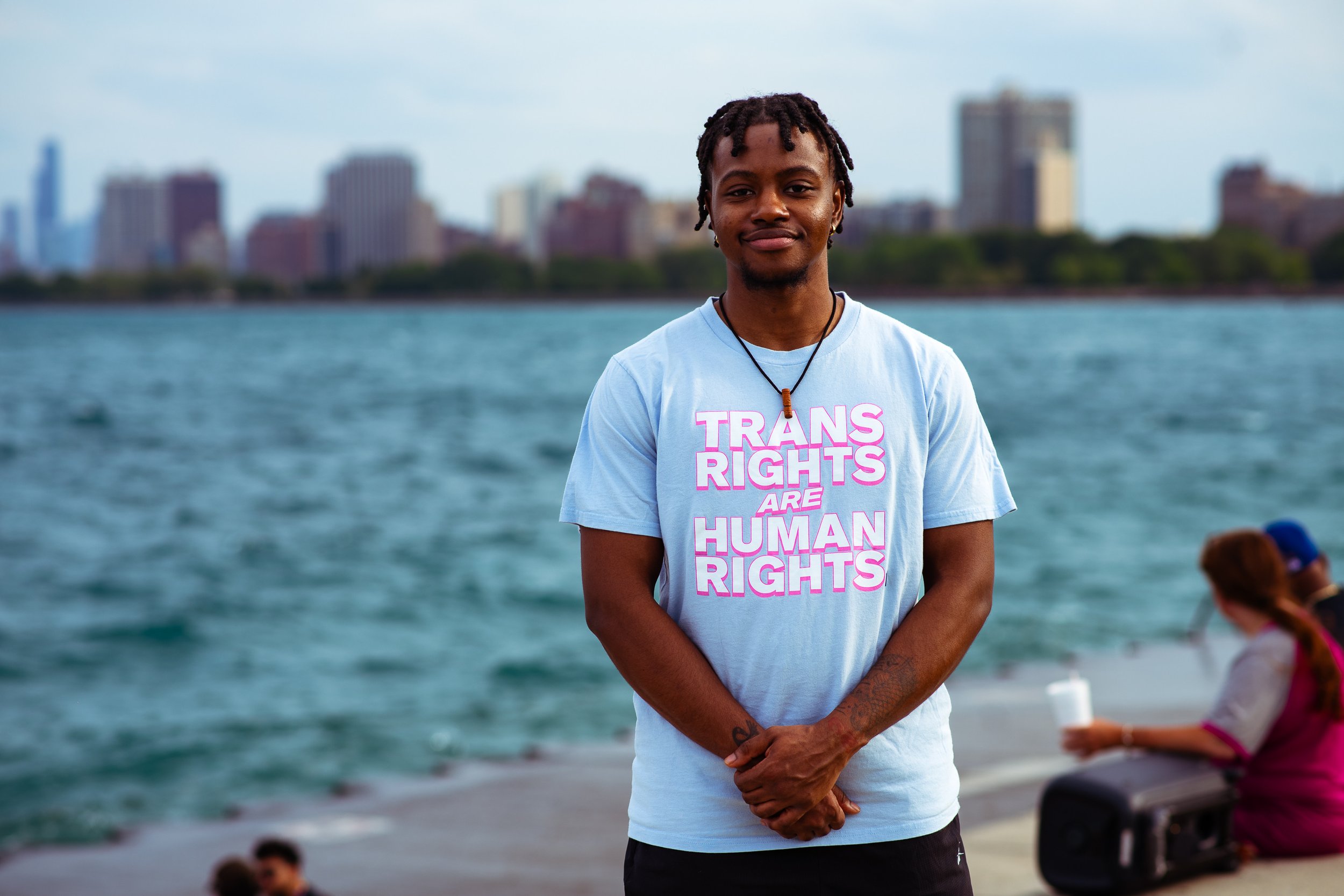 A person standing on a waterfront with a city skyline in the background, wearing a light blue T-shirt that reads 'TRANS RIGHTS ARE HUMAN RIGHTS' and a necklace with a wooden pendant.