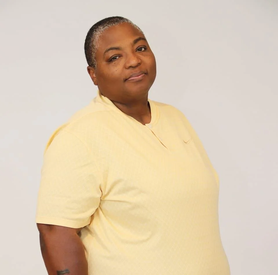 A woman with short gray hair and dark skin wearing a light yellow shirt, standing against a plain white background.