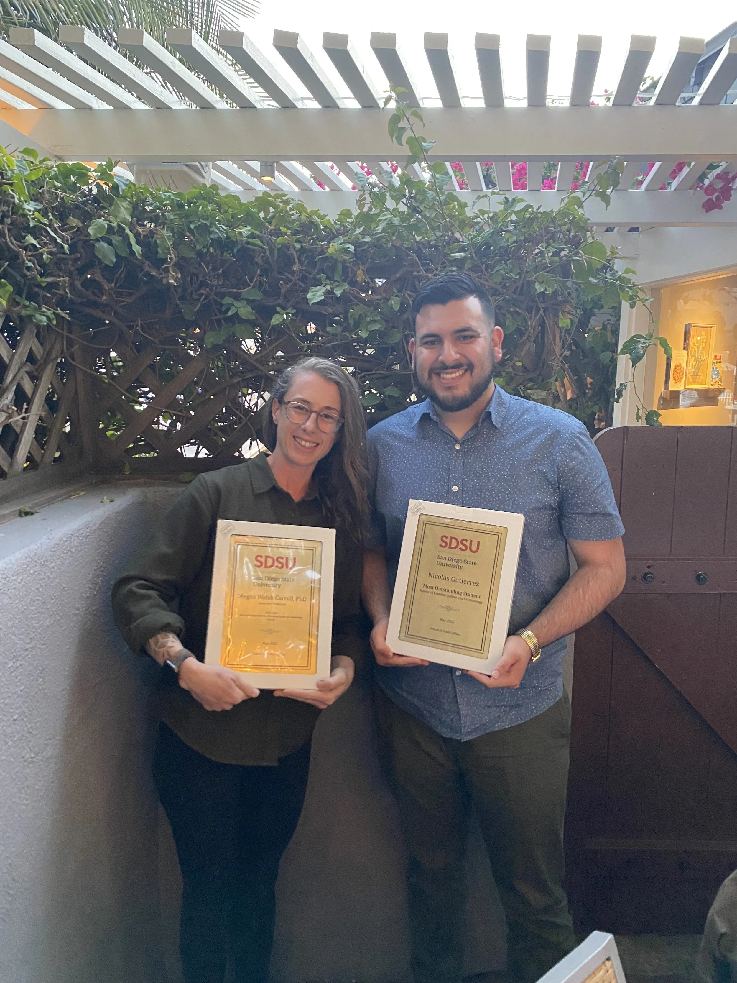 Two people smiling and holding framed certificates, standing outdoors in a setting with greenery and decorative fencing.