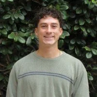 A young man with curly dark hair smiling, wearing a light green sweater, standing outdoors in front of green foliage.