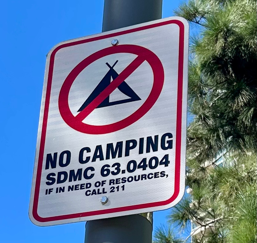 A sign indicating no camping, with a red circle and slash over a tent symbol, and text that says 'No Camping SDMC 63.0404. If in need of resources, call 211,' attached to a pole outdoors with trees and a blue sky in the background.