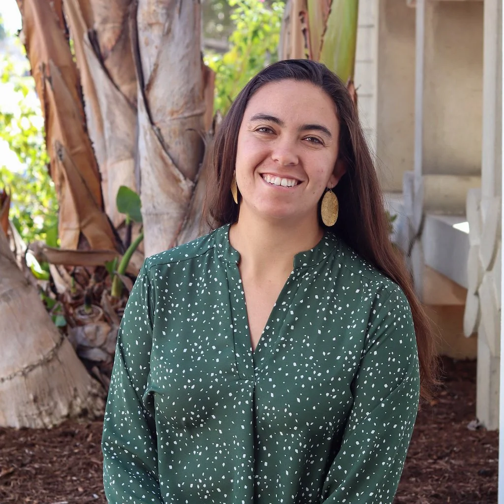 A woman with long dark hair smiling outdoors near a large tree with peeling bark and green foliage, wearing a green blouse with white speckles and gold earrings.